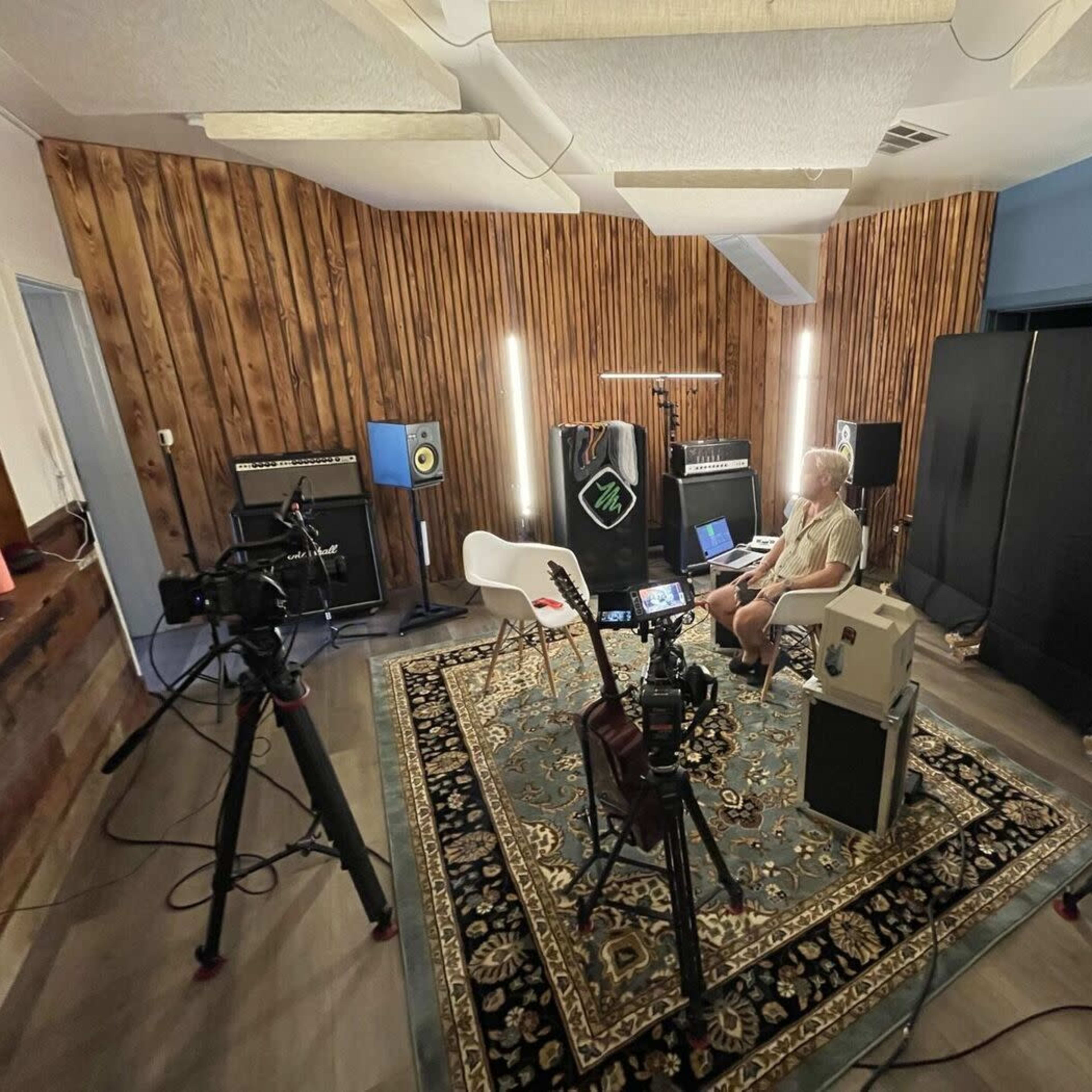 A recording studio with a person sitting in a chair surrounded by cameras, audio equipment, and acoustic panels.