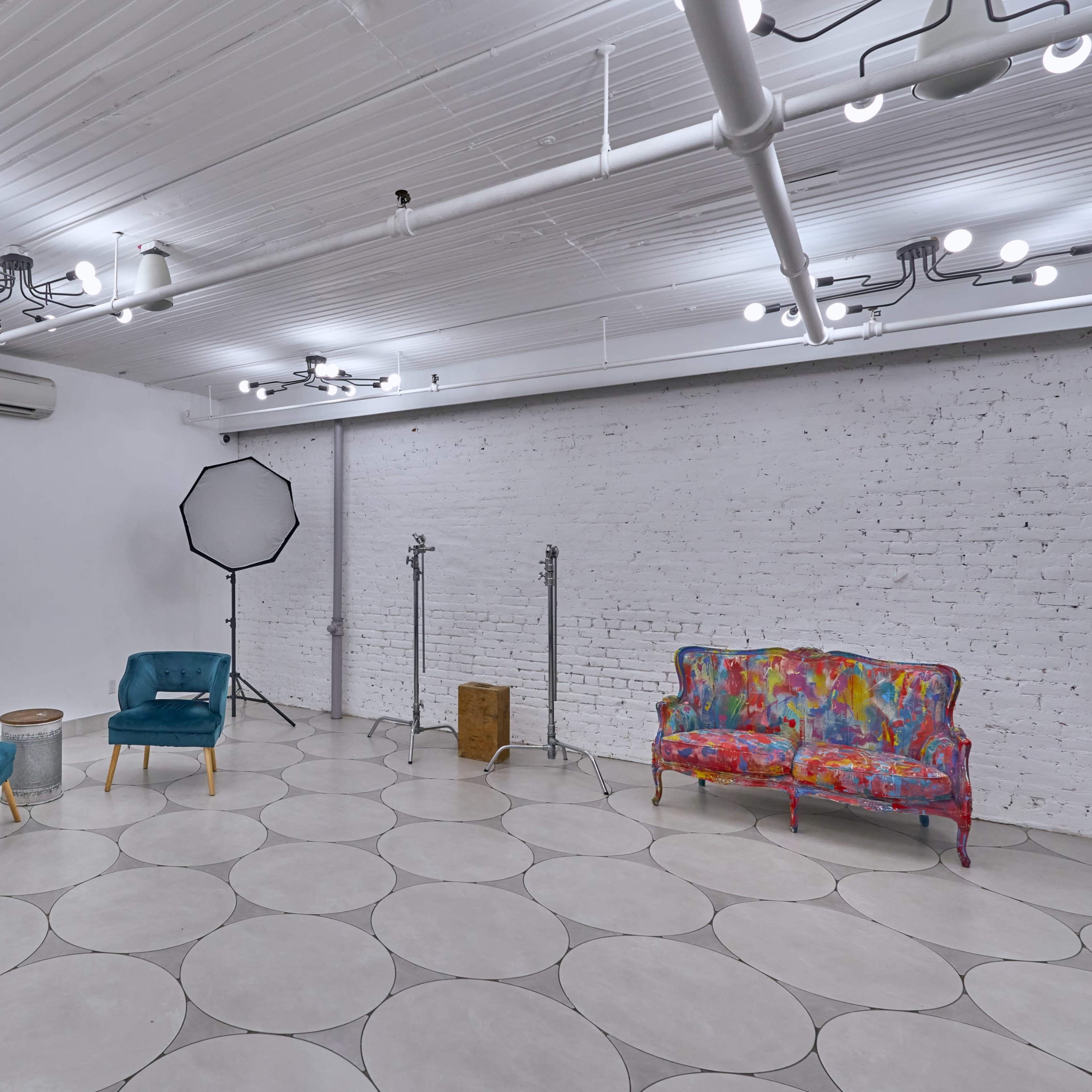 A spacious, brightly lit room with two teal chairs, a colorful patterned couch, and a photo backdrop setup, all arranged on a circular tiled floor.