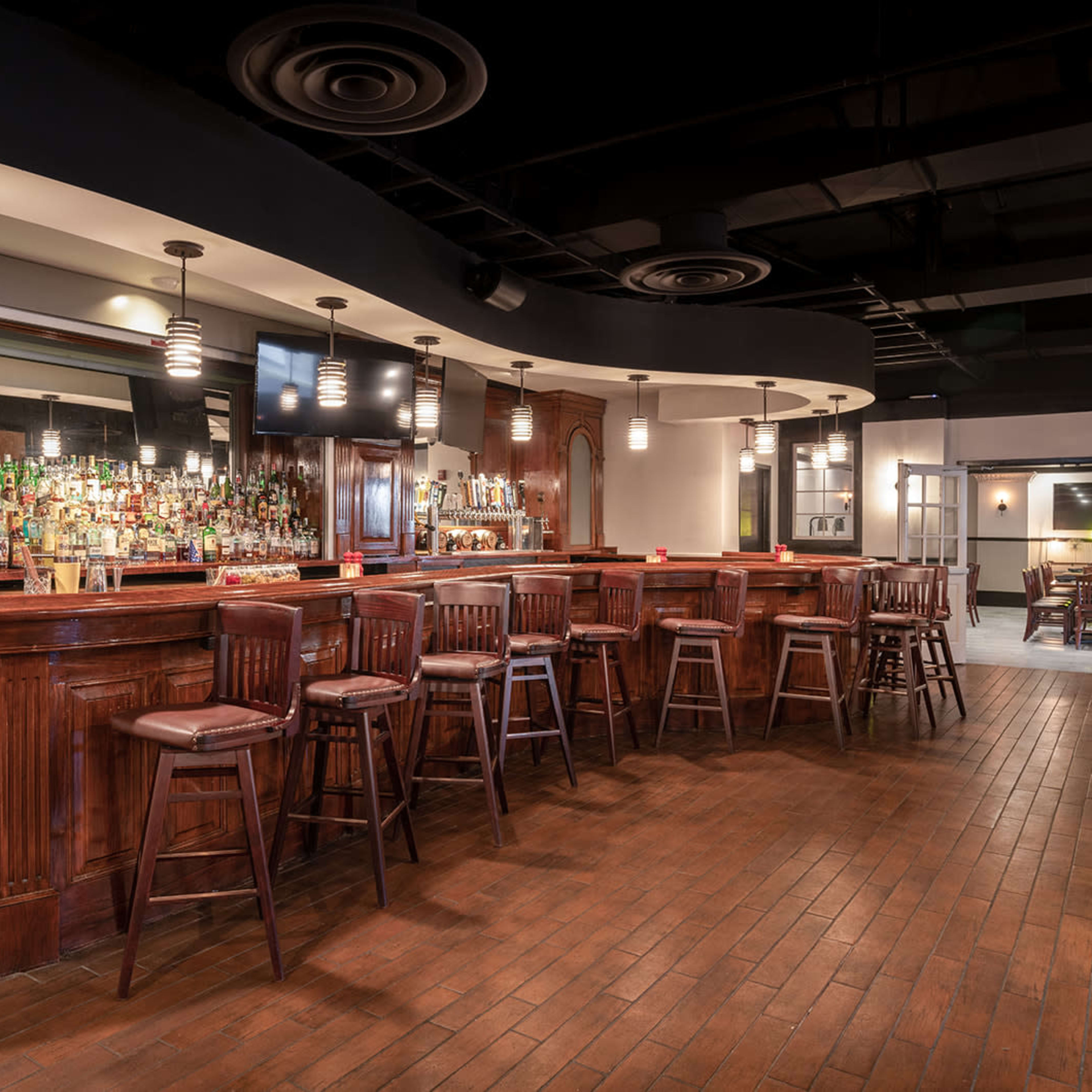 A wooden bar area with high stools, a mirrored back wall filled with bottles, and pendant lighting in a modern interior space.