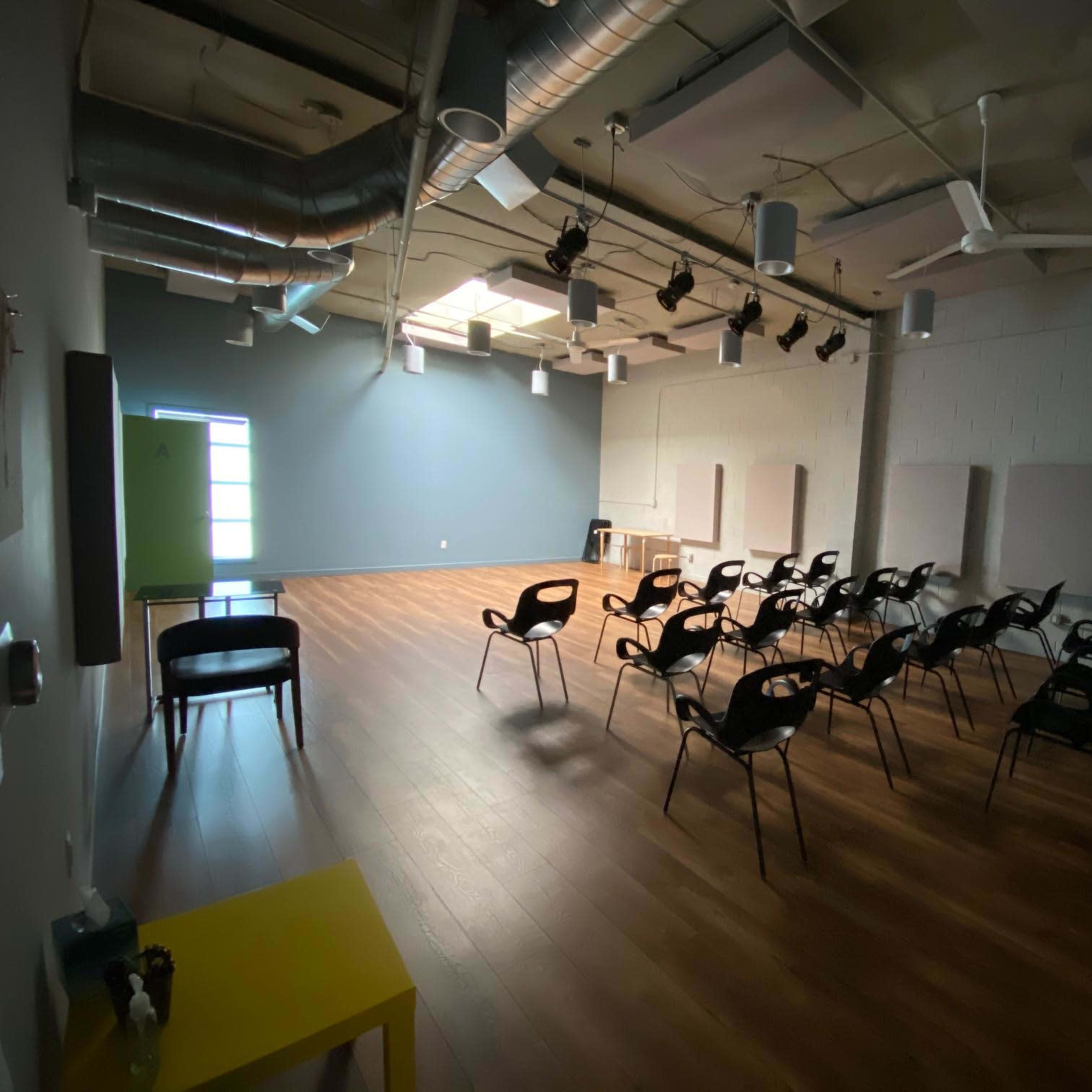 A sparsely furnished room featuring wooden floors, rows of black chairs, and track lighting, with a large window on one wall.