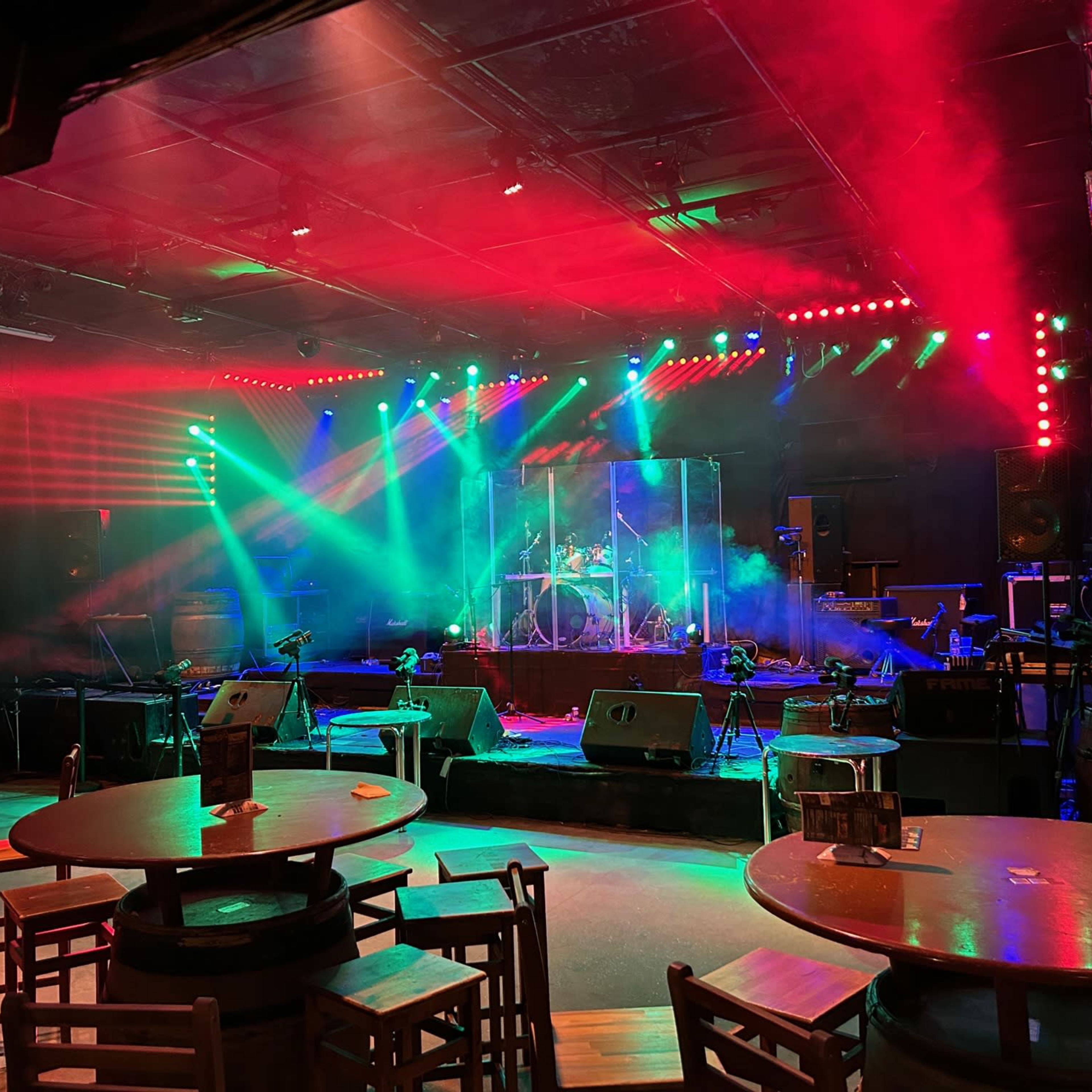 A stage setup with bright multicolored lights, fog effects, and musical instruments prepared for a performance, surrounded by empty tables and chairs.