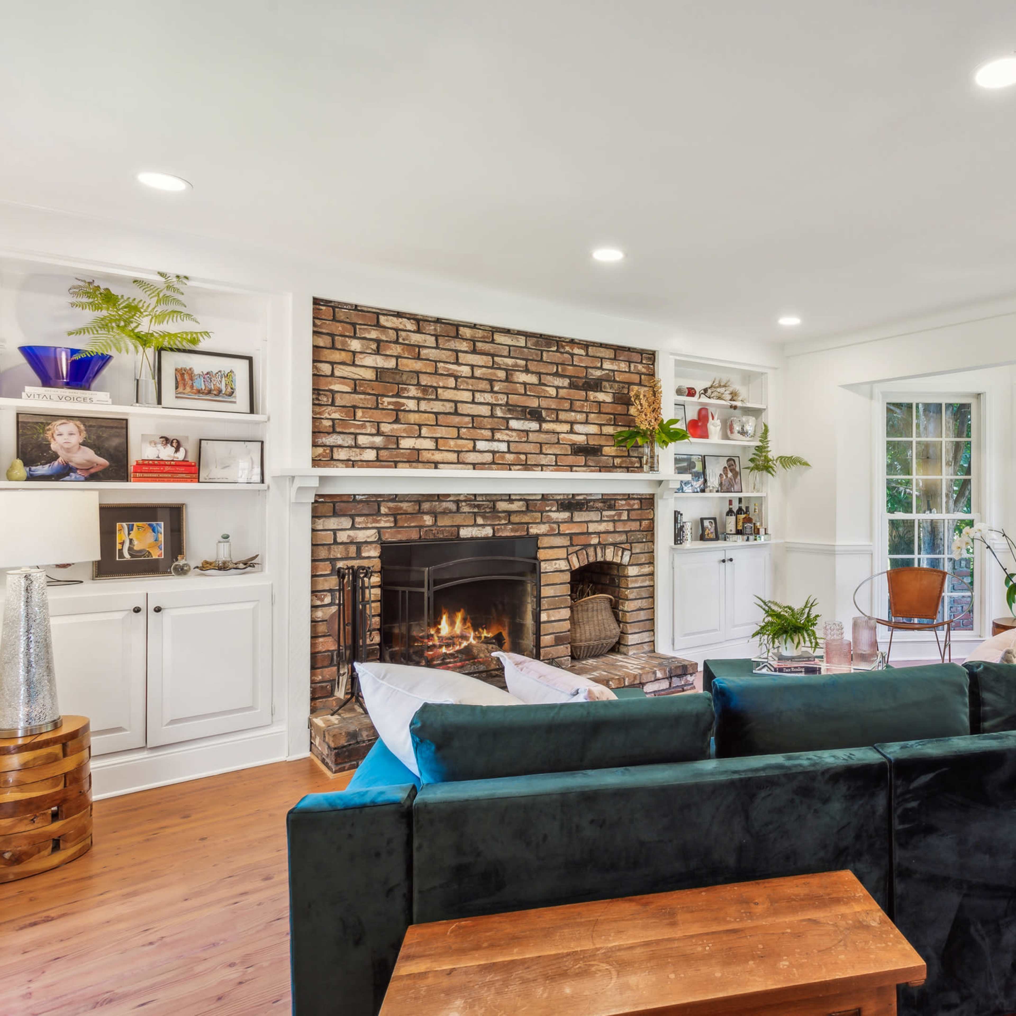 The living room features a brick fireplace with built-in shelves, a dark sofa, and large windows that overlook a garden.