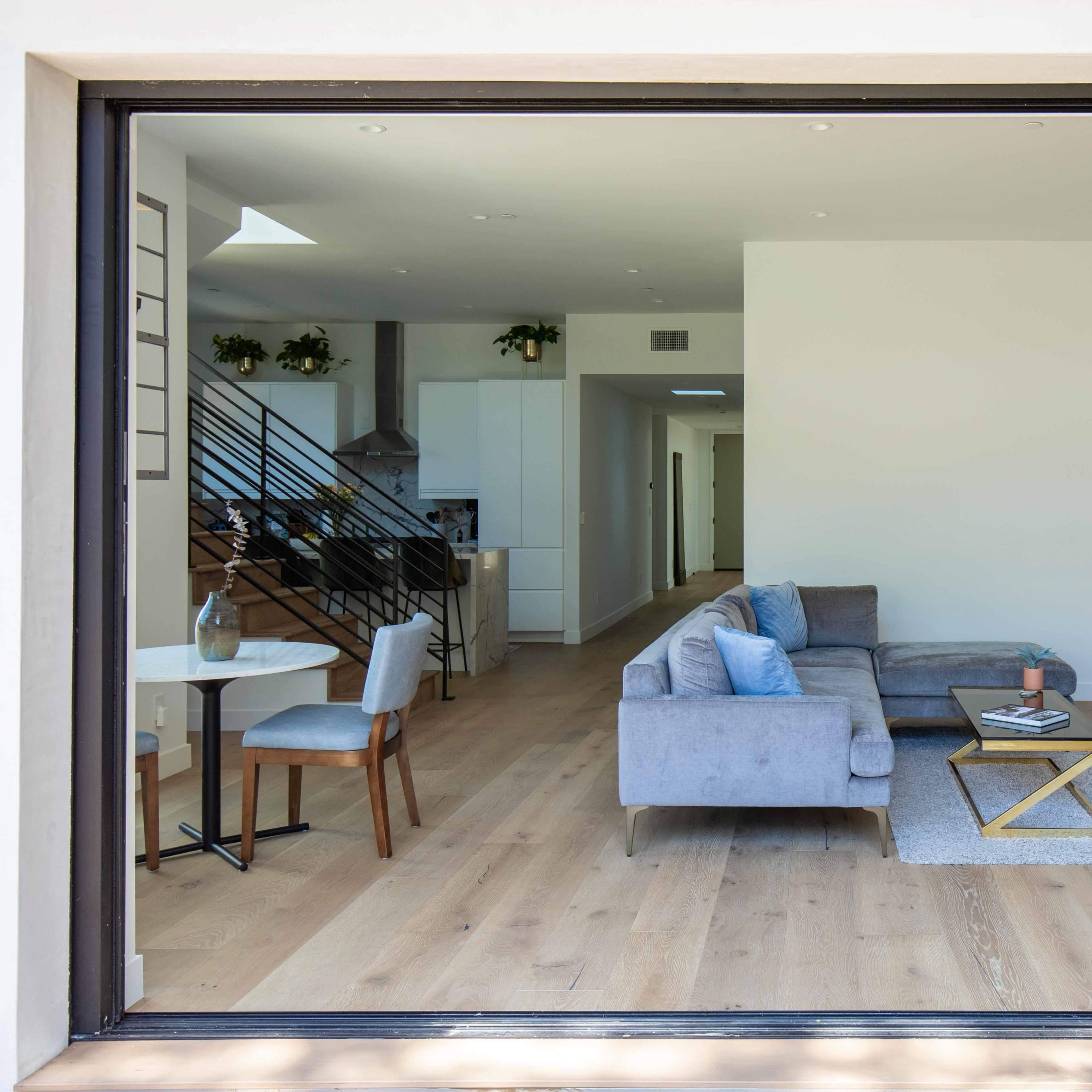 A large sliding glass door reveals a modern living space with a sectional sofa, a coffee table on a rug, and a dining area with a small table.