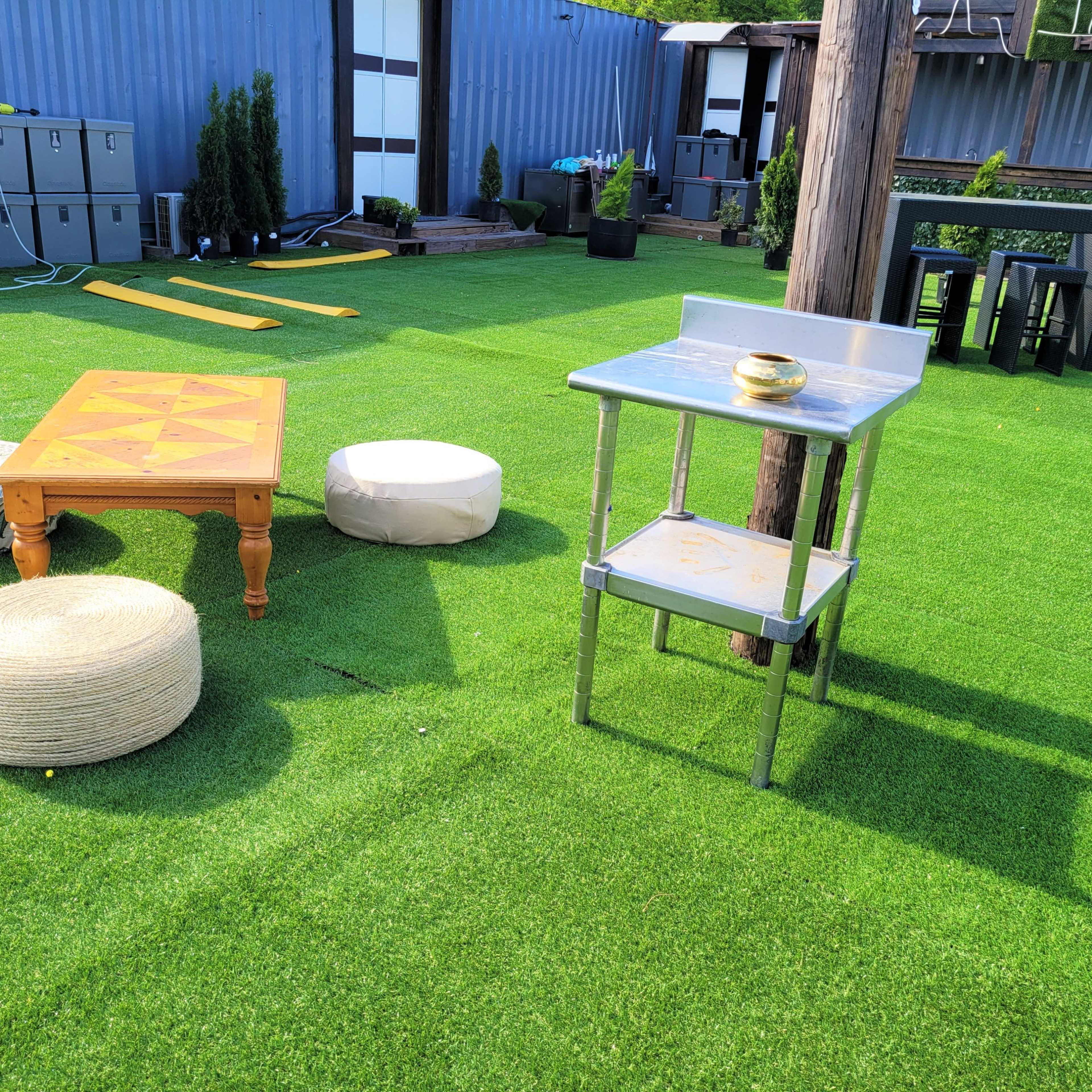 The scene features a green artificial grass area with a wooden table, round cushions, and a metal kitchen prep table.