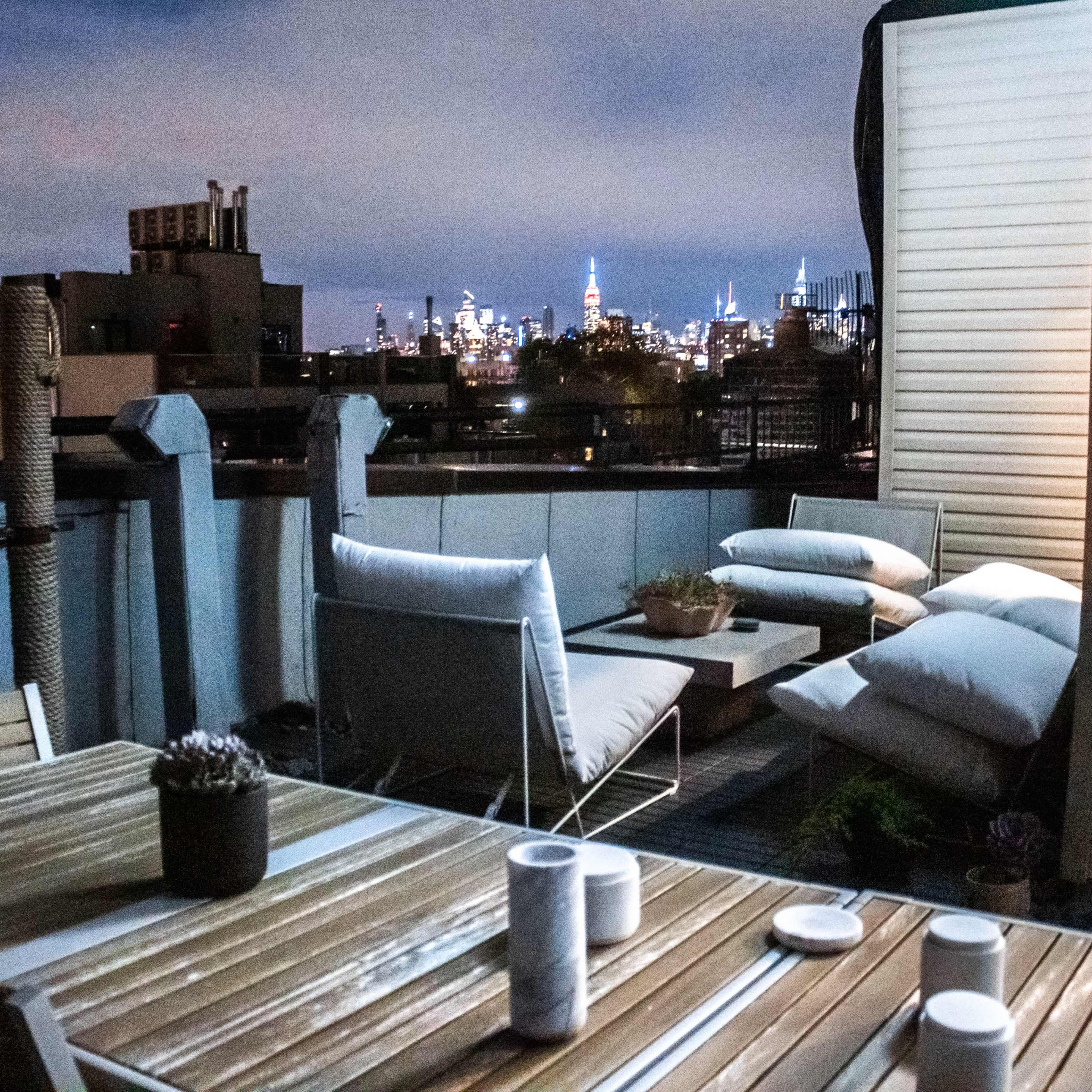 A rooftop terrace features a wooden table with candles and minimal seating, overlooking a skyline with illuminated buildings at night.