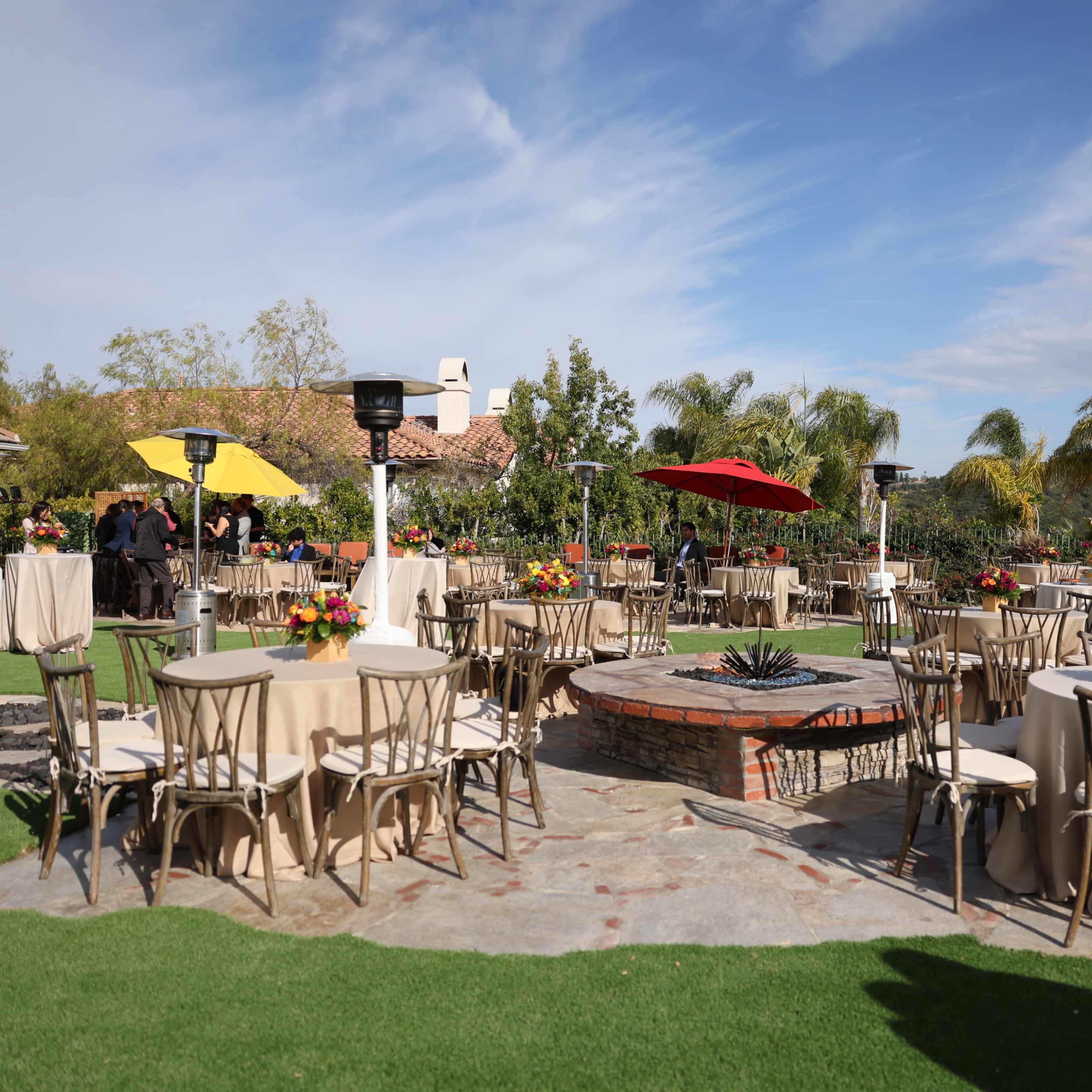 A landscaped outdoor venue features round tables with beige tablecloths, surrounded by greenery and decorative flowers, with a fire pit at the center.
