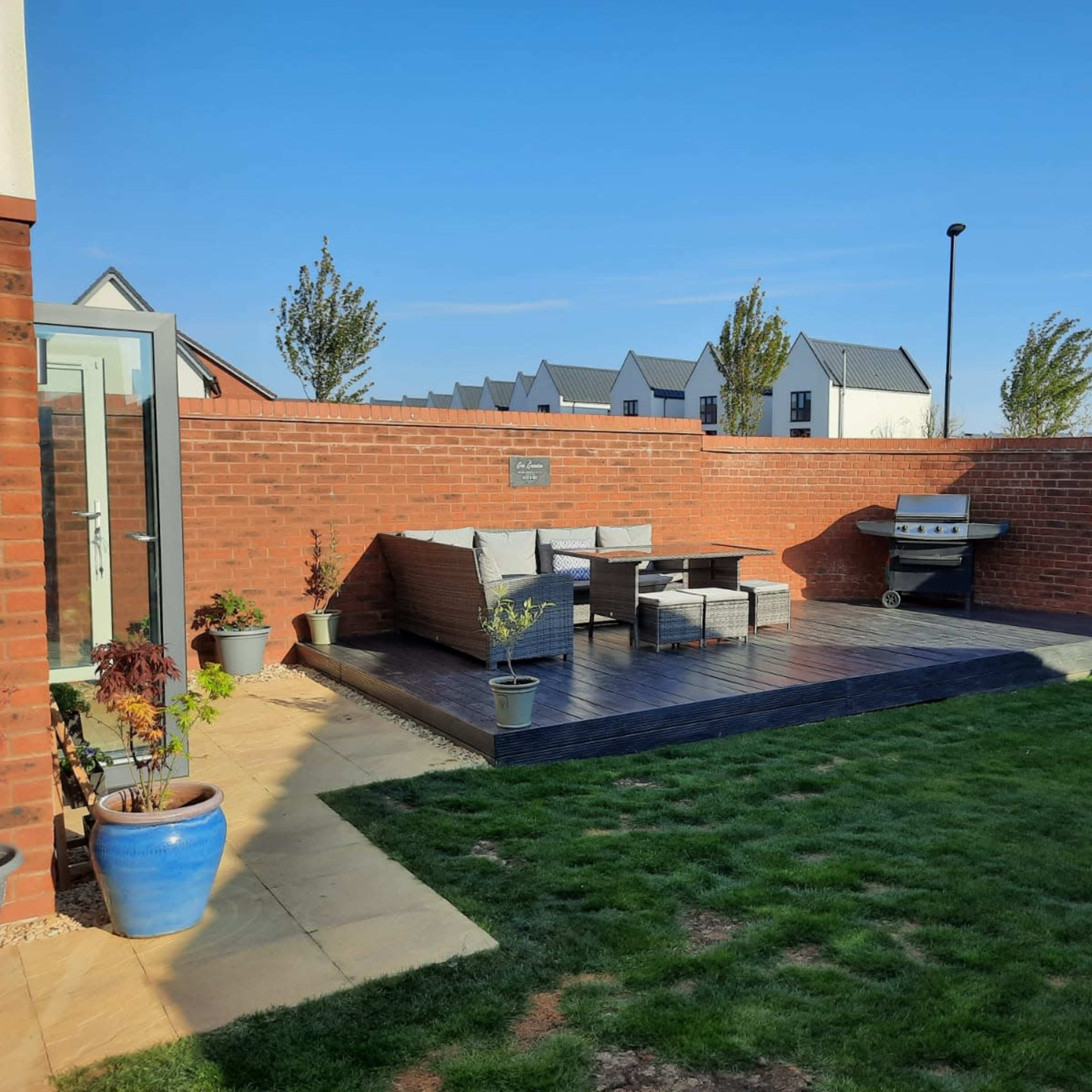 A landscaped backyard featuring a wooden deck with outdoor seating, a grill, and a grassy area surrounded by brick walls.
