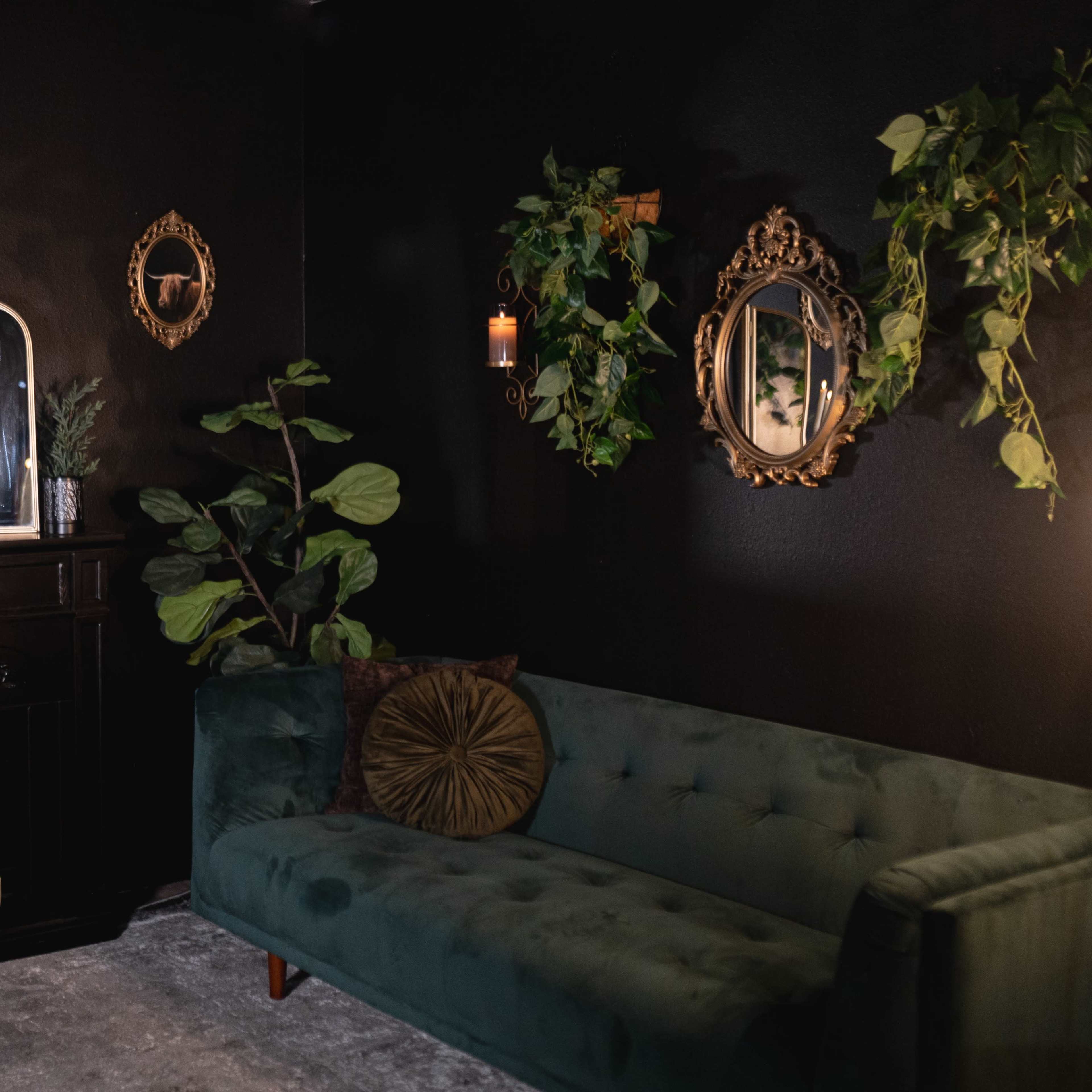 A dark room featuring a green velvet sofa, a wooden cabinet with candles, decorative mirrors, and wall decor including a skull and plants.