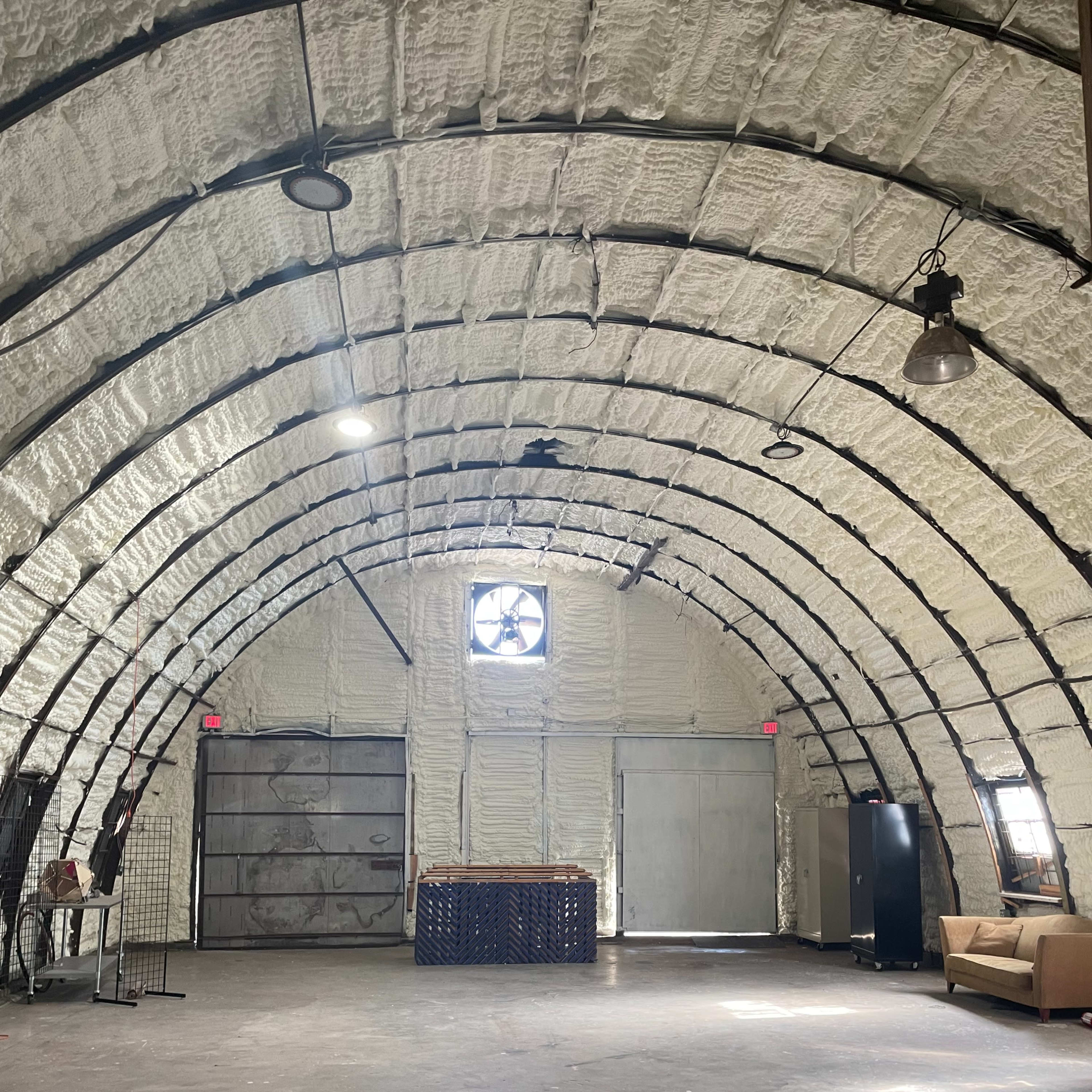 The interior of a large, arched building features exposed insulation, a fan in the window, and a couch against one wall.