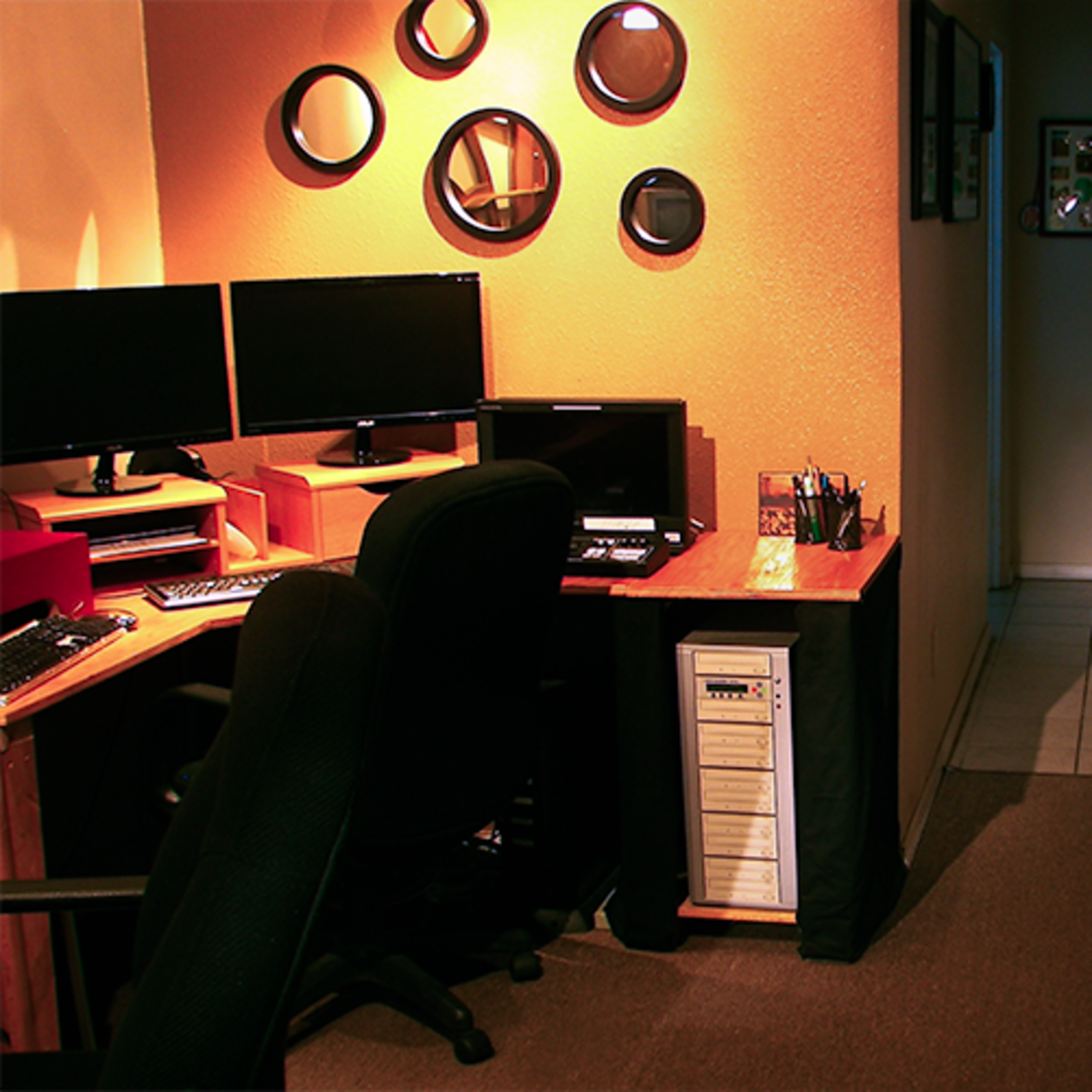 A small office space with two computer monitors, a desk with a CPU, and a seating area featuring red chairs, leading to a hallway and a kitchen area in the background.
