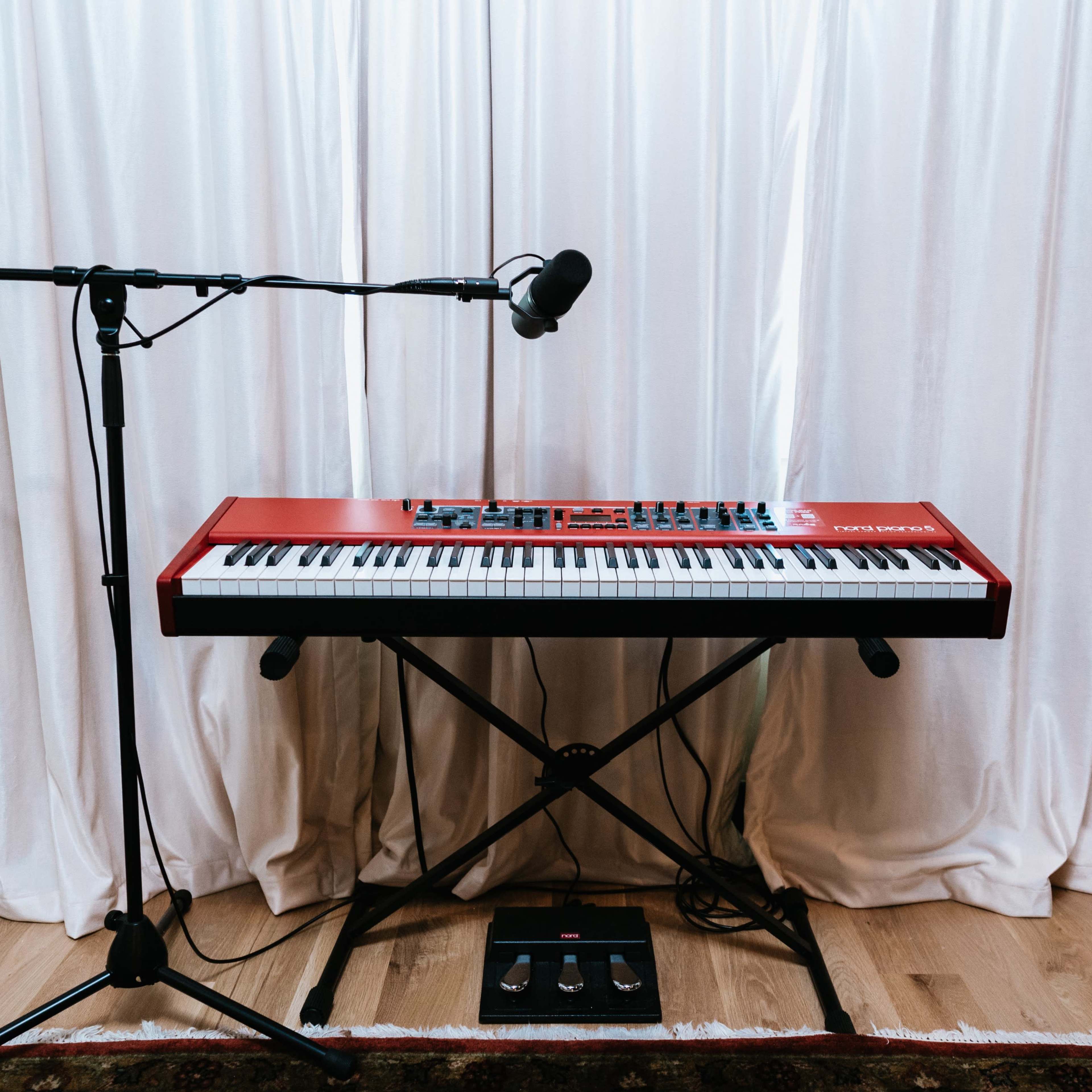 A red stage piano with a microphone stand and foot pedals is positioned in front of a white curtain.