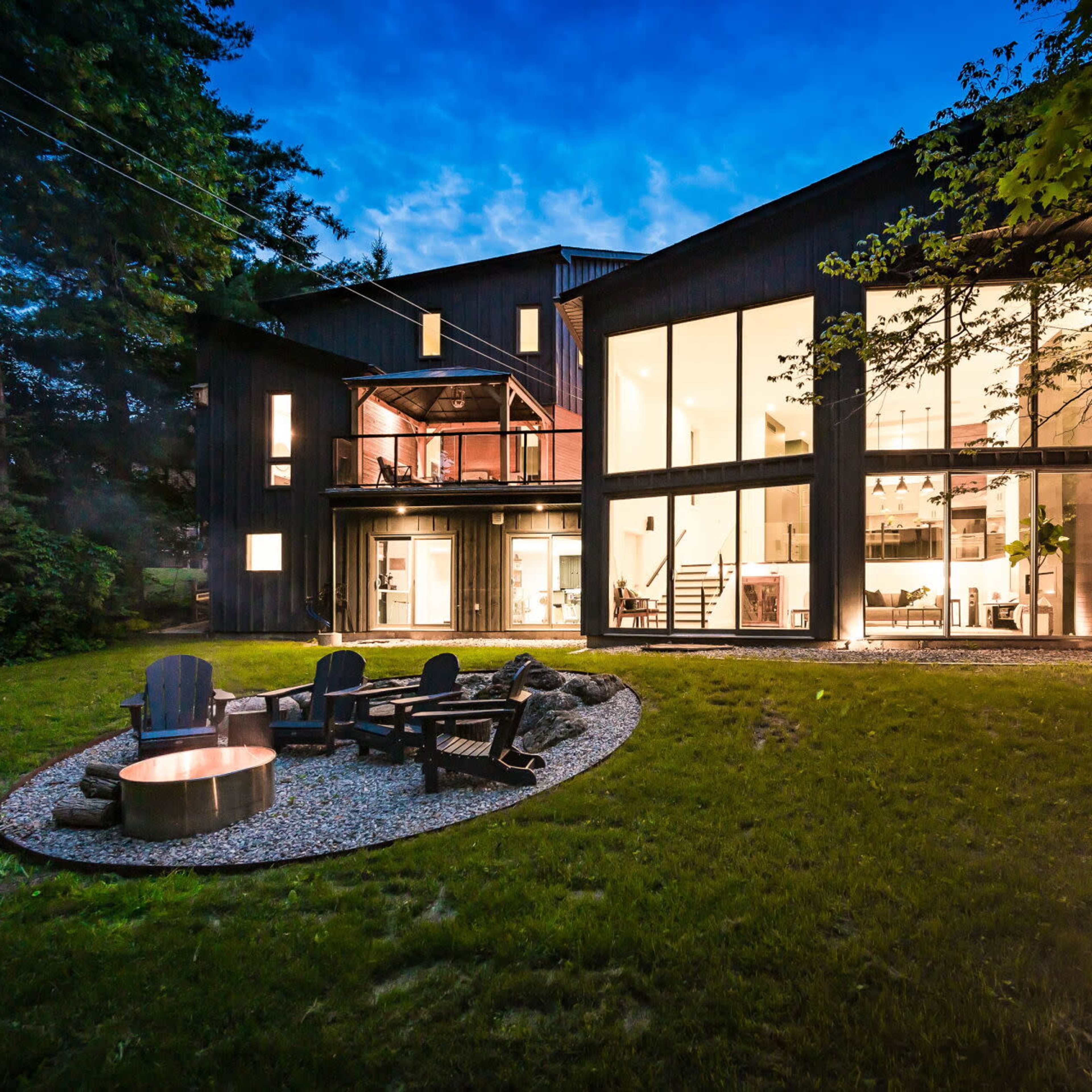 A modern two-story house with large glass windows is surrounded by trees and features a circular fire pit with wooden chairs in the backyard.
