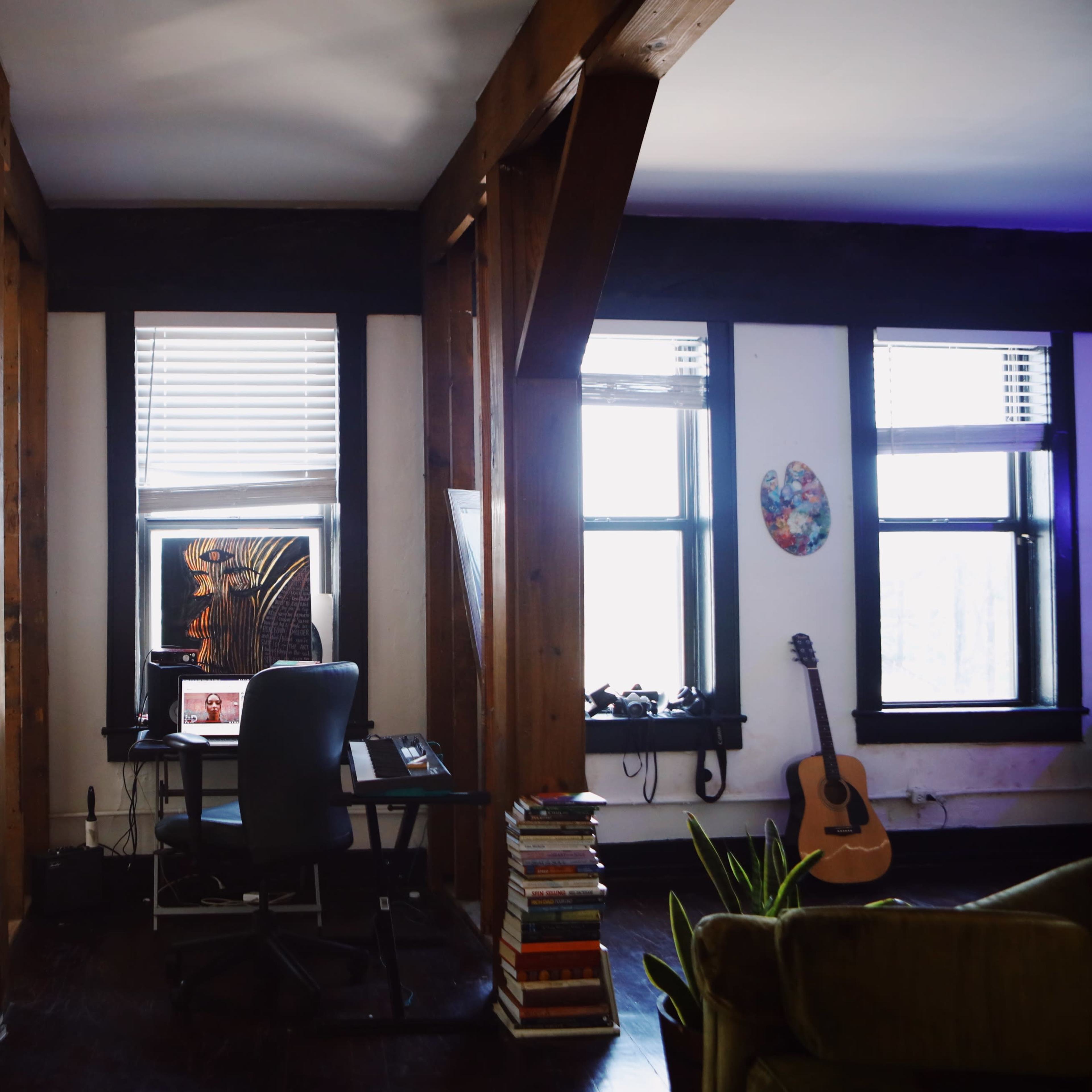 An interior space featuring a desk with a computer, stacked books, a guitar, and large windows with wooden beams and artwork on the walls.