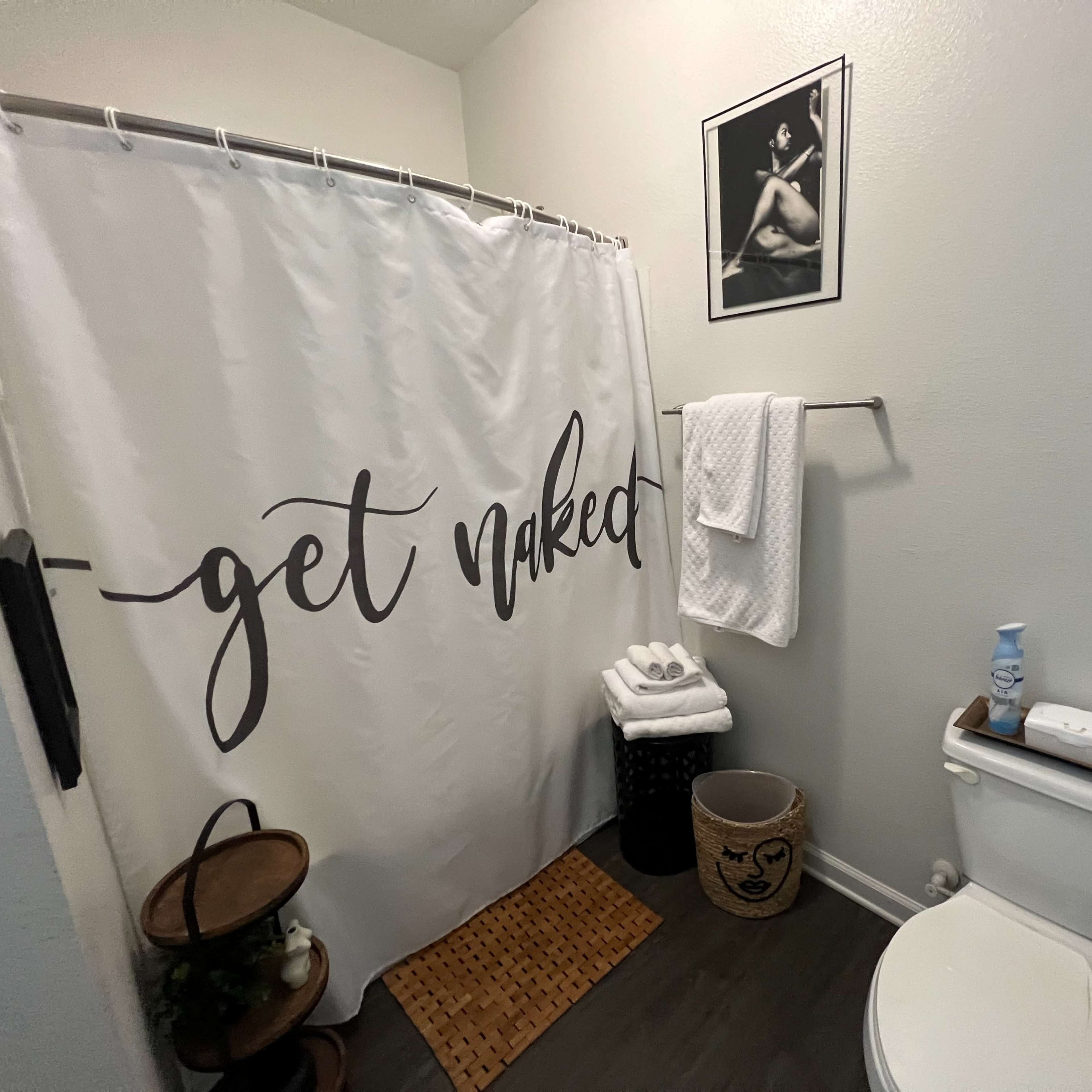 A bathroom with a shower curtain that reads "get naked," a toilet, neatly stacked white towels, and a small wooden stool beside a woven basket.