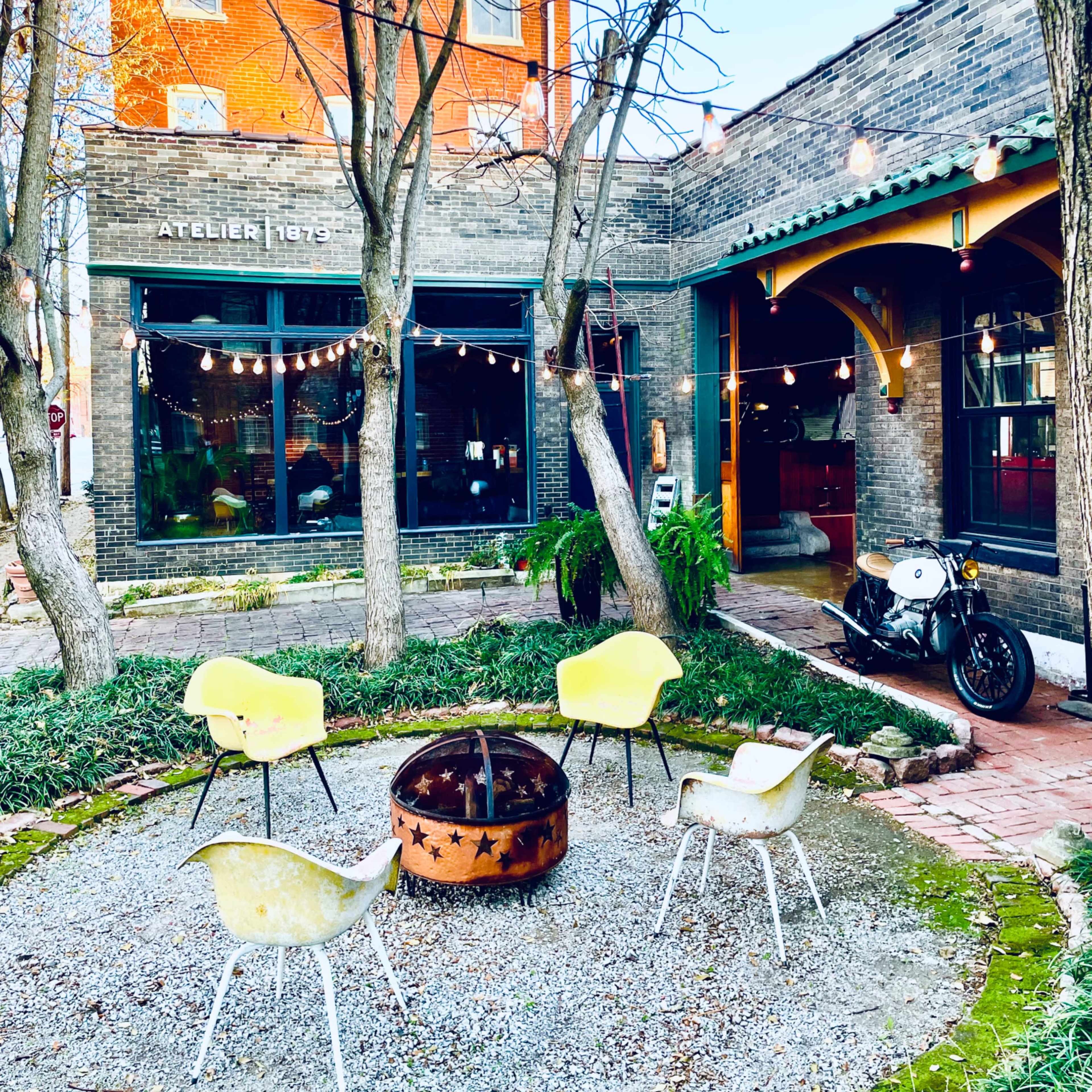 A circular gravel seating area with four yellow chairs arranged around a fire pit, bordered by trees and adjacent to a brick building with large windows and string lights.