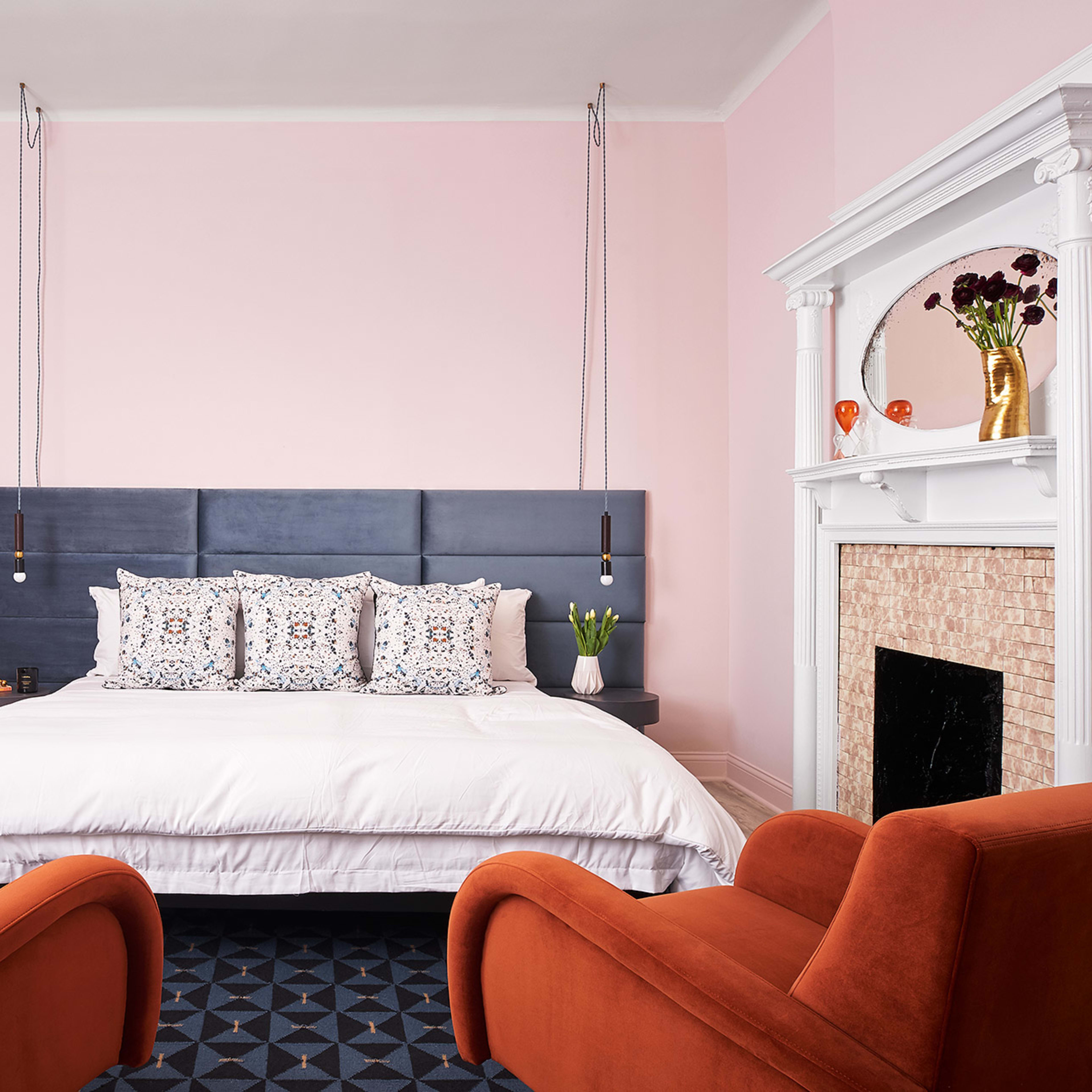A modern bedroom with a large bed flanked by two orange chairs, featuring a pink wall and a decorative fireplace.