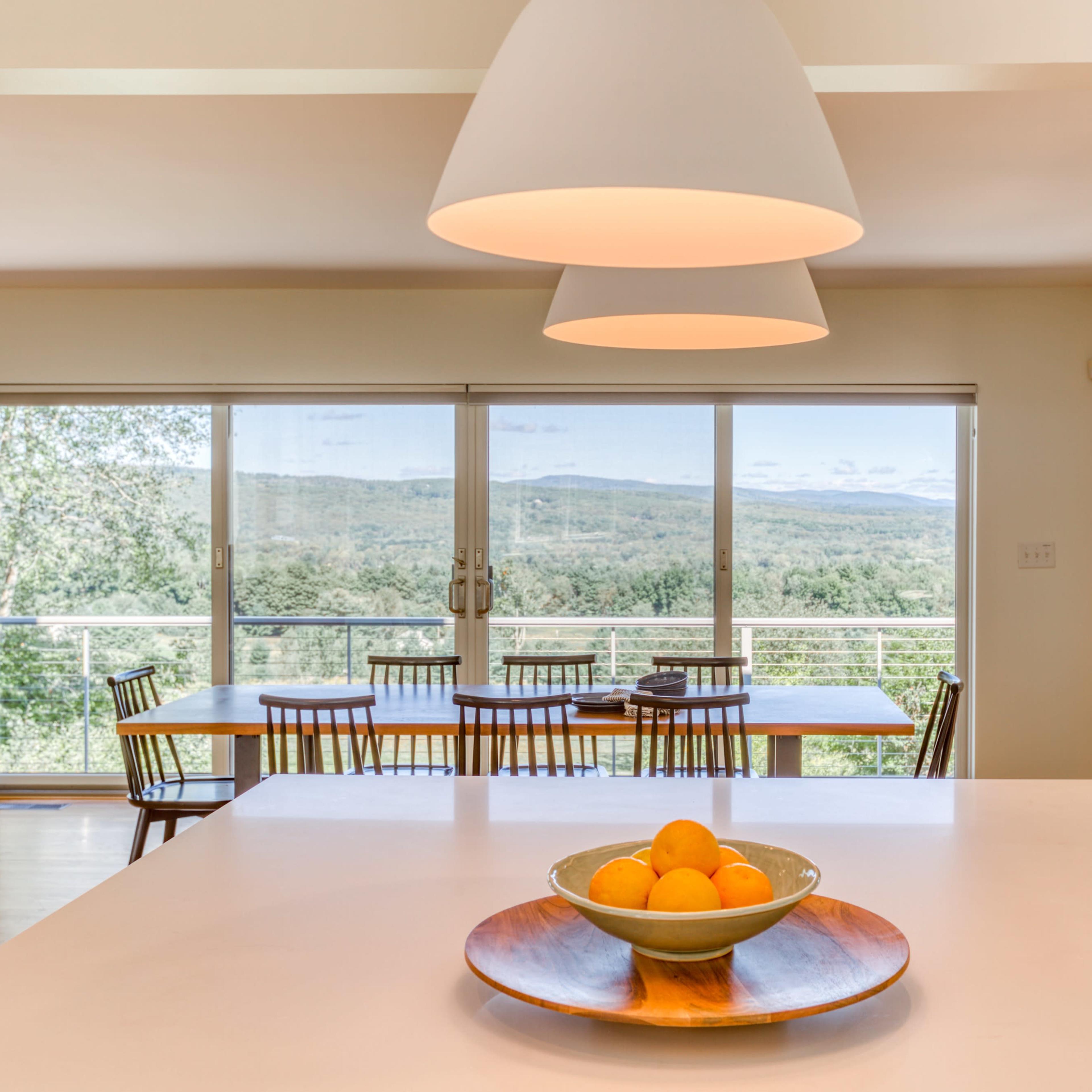 A modern dining area features a white table with a bowl of oranges, large windows offering a view of rolling hills, and a set of wooden chairs arranged around a dining table.