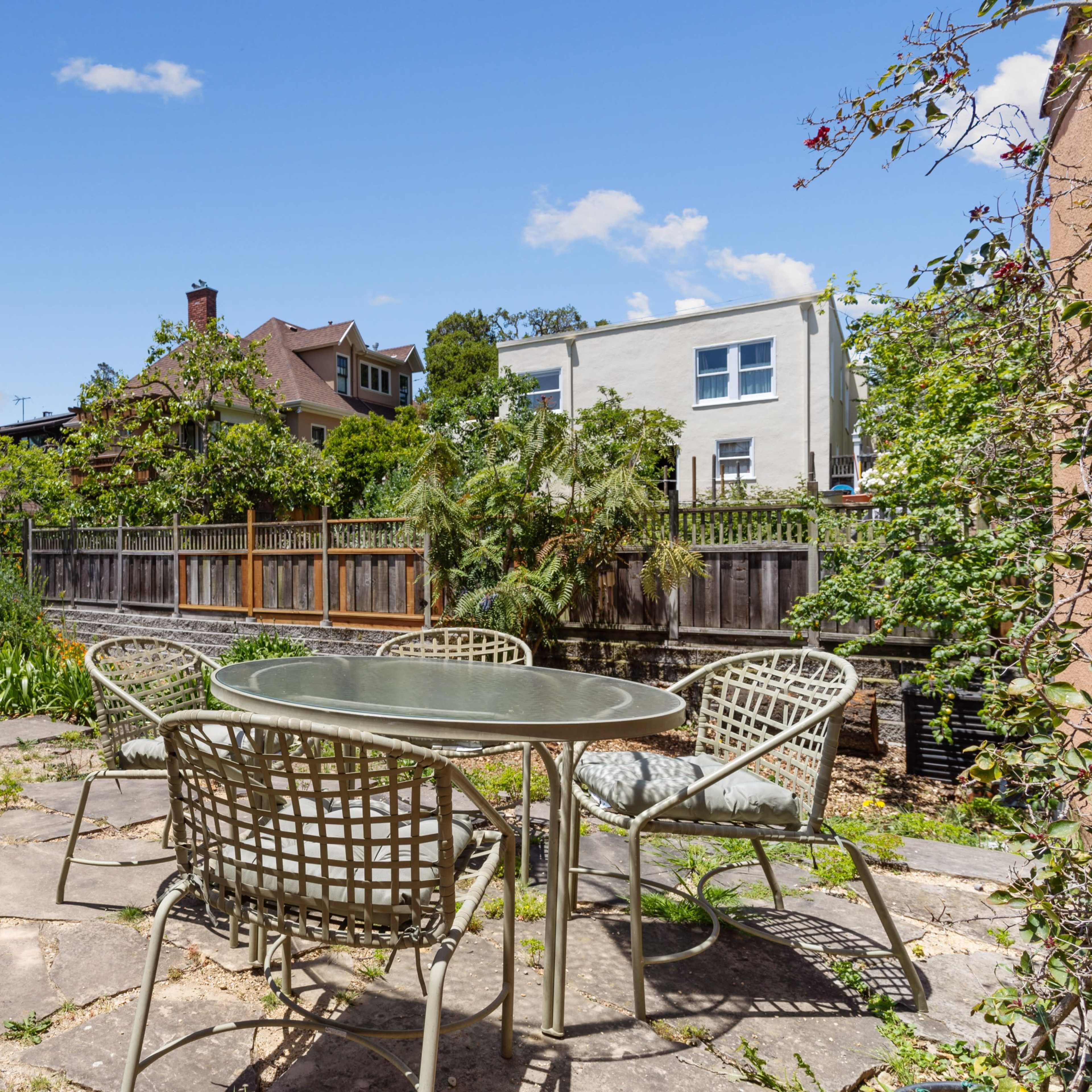 A stone patio area with a glass-top table and four chairs, surrounded by greenery and residential buildings in the background.