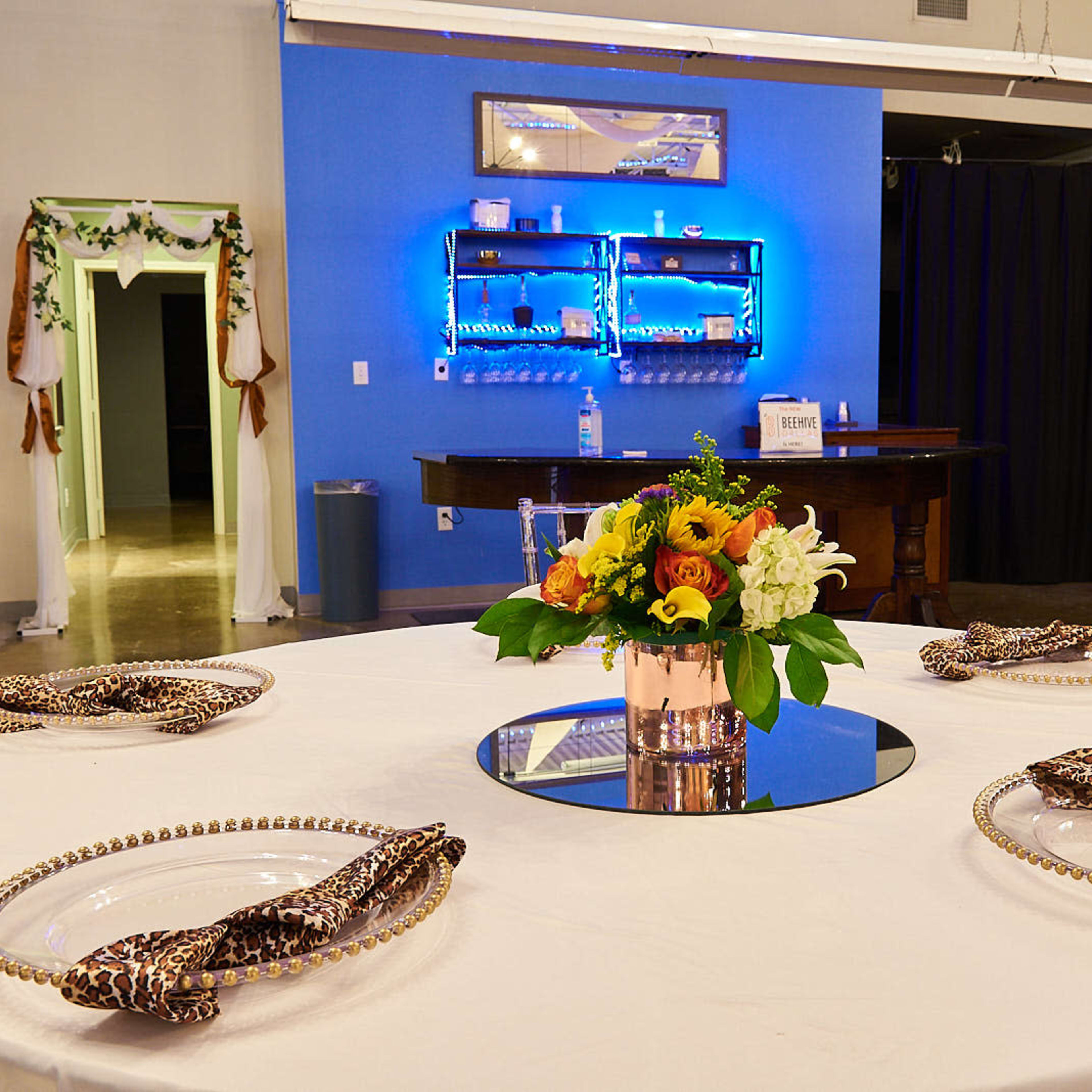 A round dining table is set with elegant plates and a floral centerpiece in a well-lit room featuring blue and dark curtains in the background.