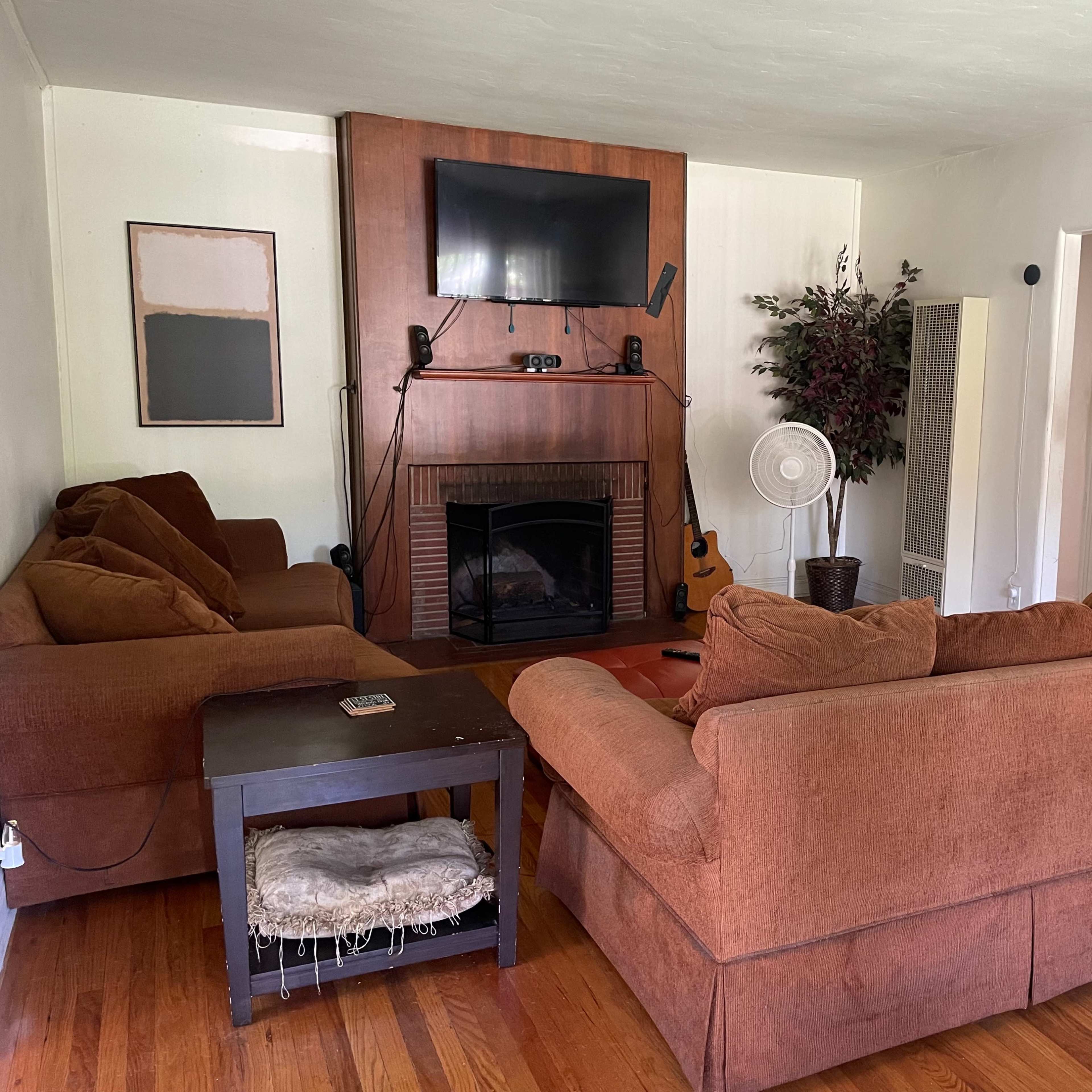 A living room with a sofa set, a coffee table, a wall-mounted TV, a fireplace, and a fan in front of a houseplant.