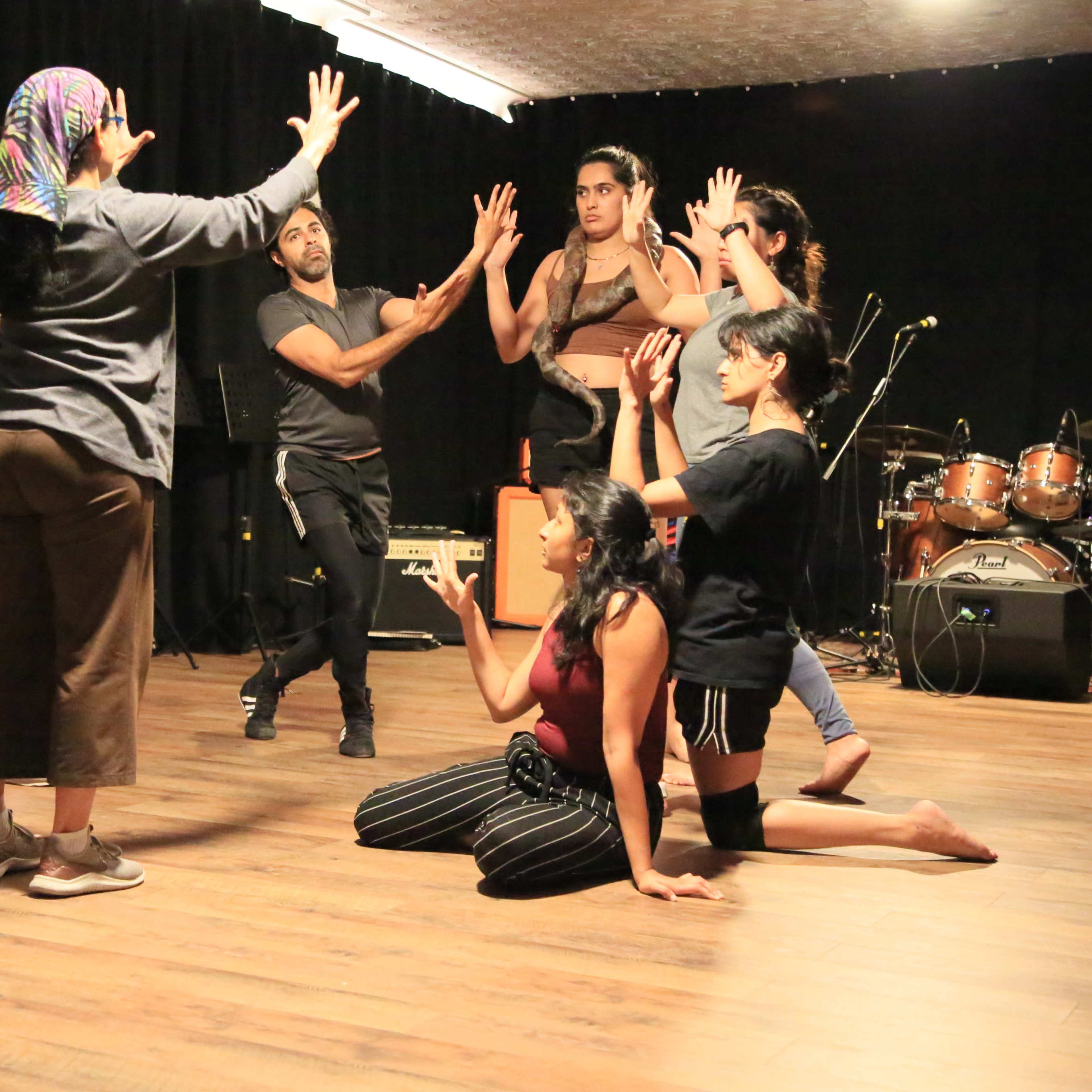 A group of performers rehearses on a wooden stage while a woman leads the session with her hands raised.