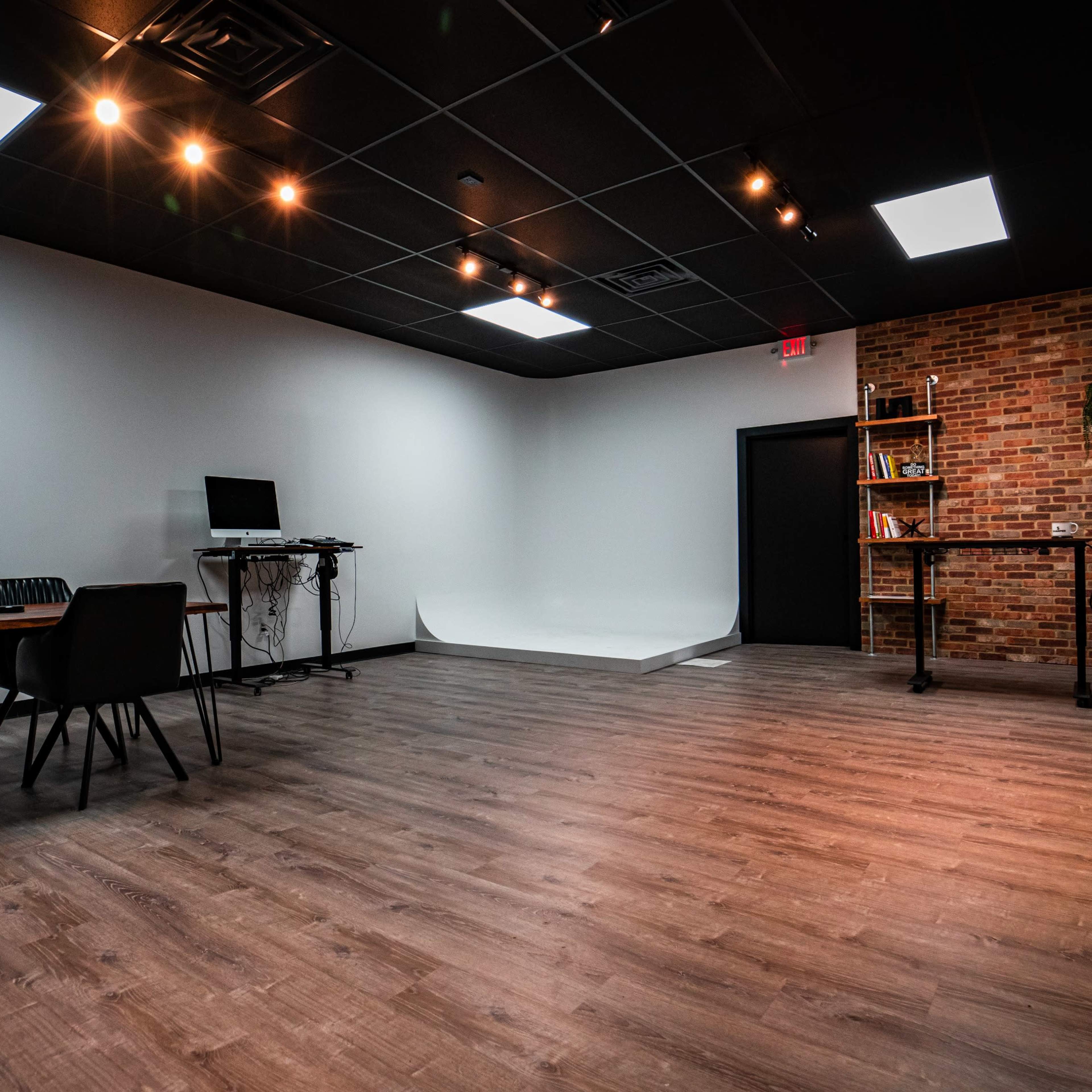 A modern, empty studio space with wooden flooring, two tables with chairs, a computer setup, and a brick wall with shelves.