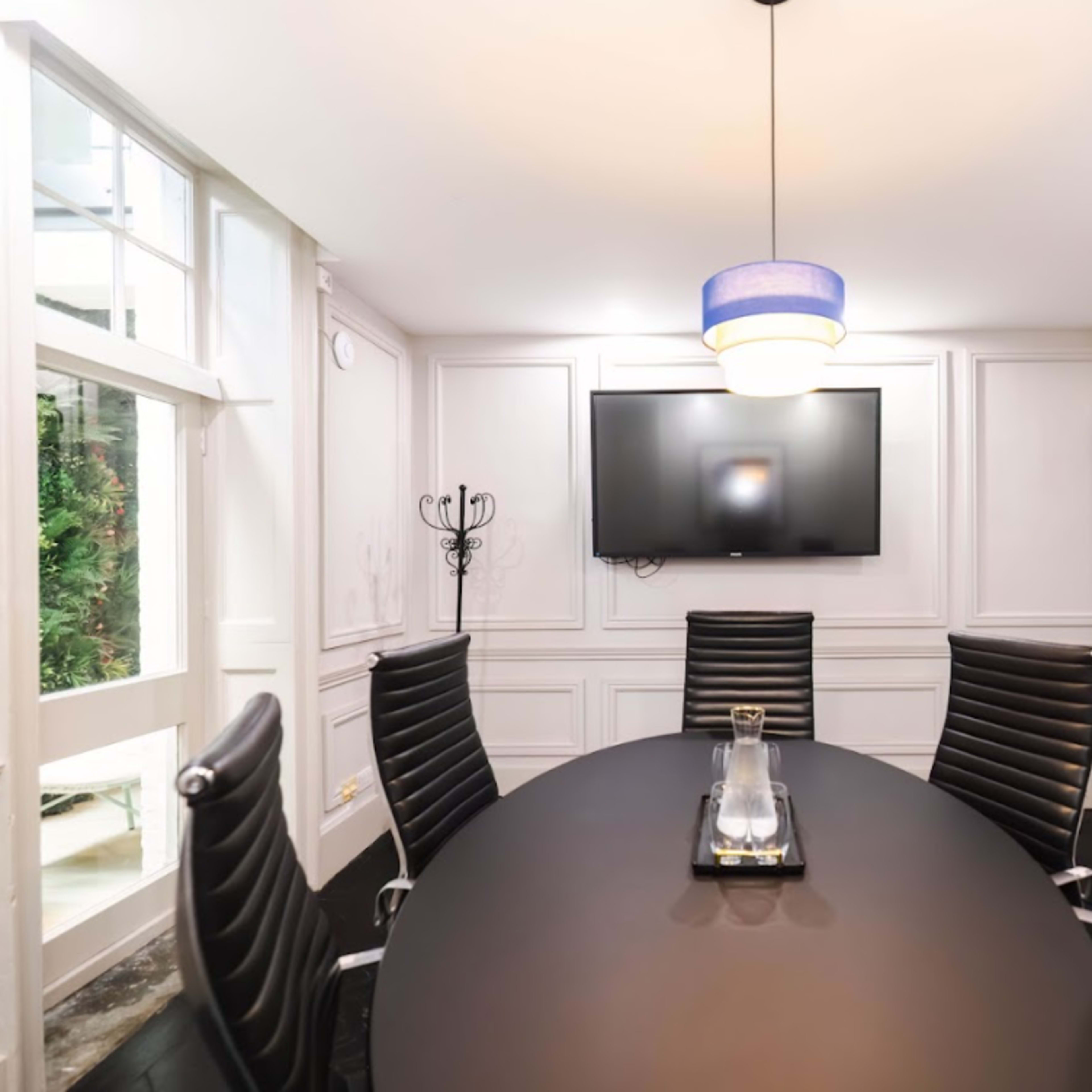 A modern conference room features a large oval table surrounded by black leather chairs, with a television mounted on a white paneled wall.