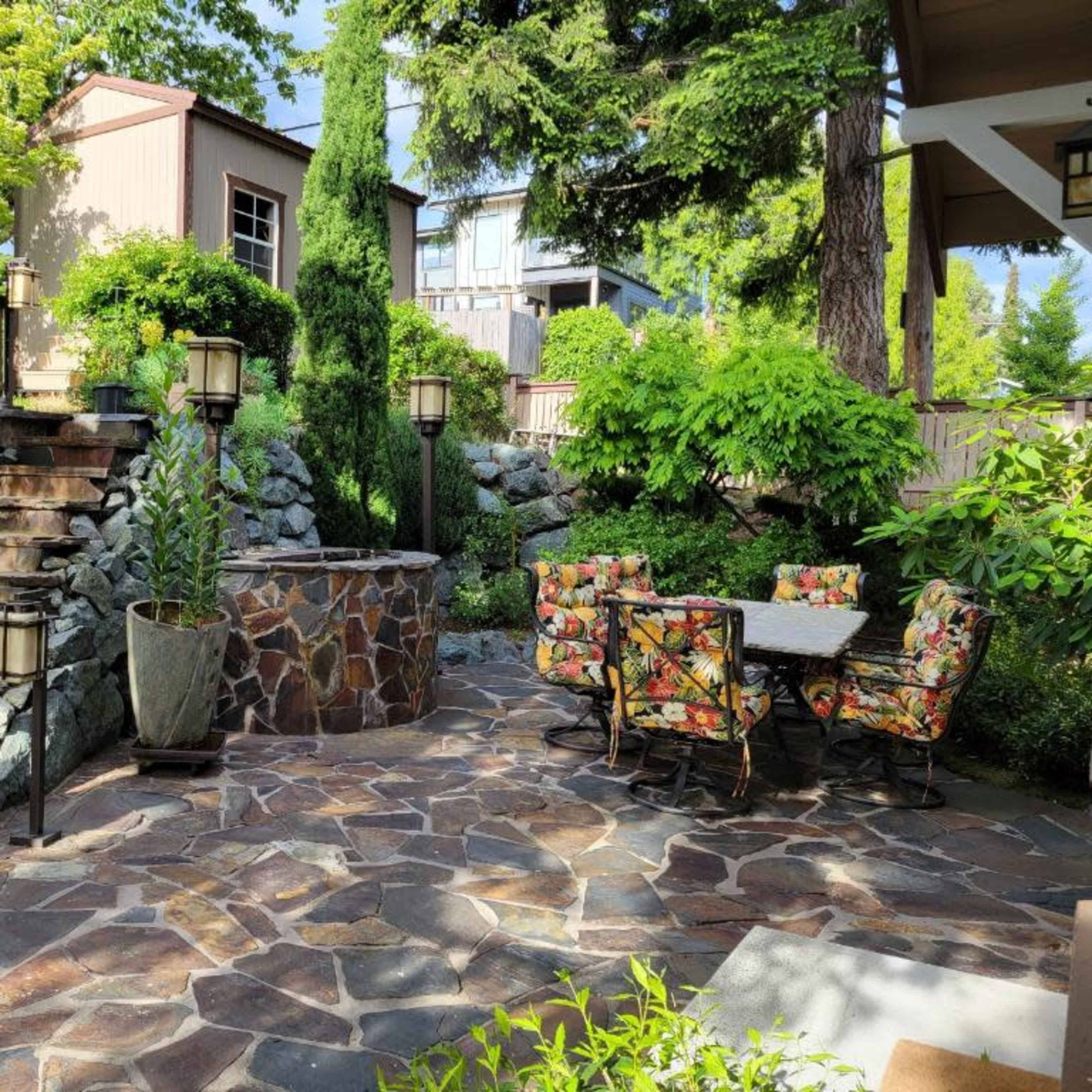 A landscaped outdoor area featuring a stone patio with a dining set surrounded by greenery and stairs leading to a higher level.