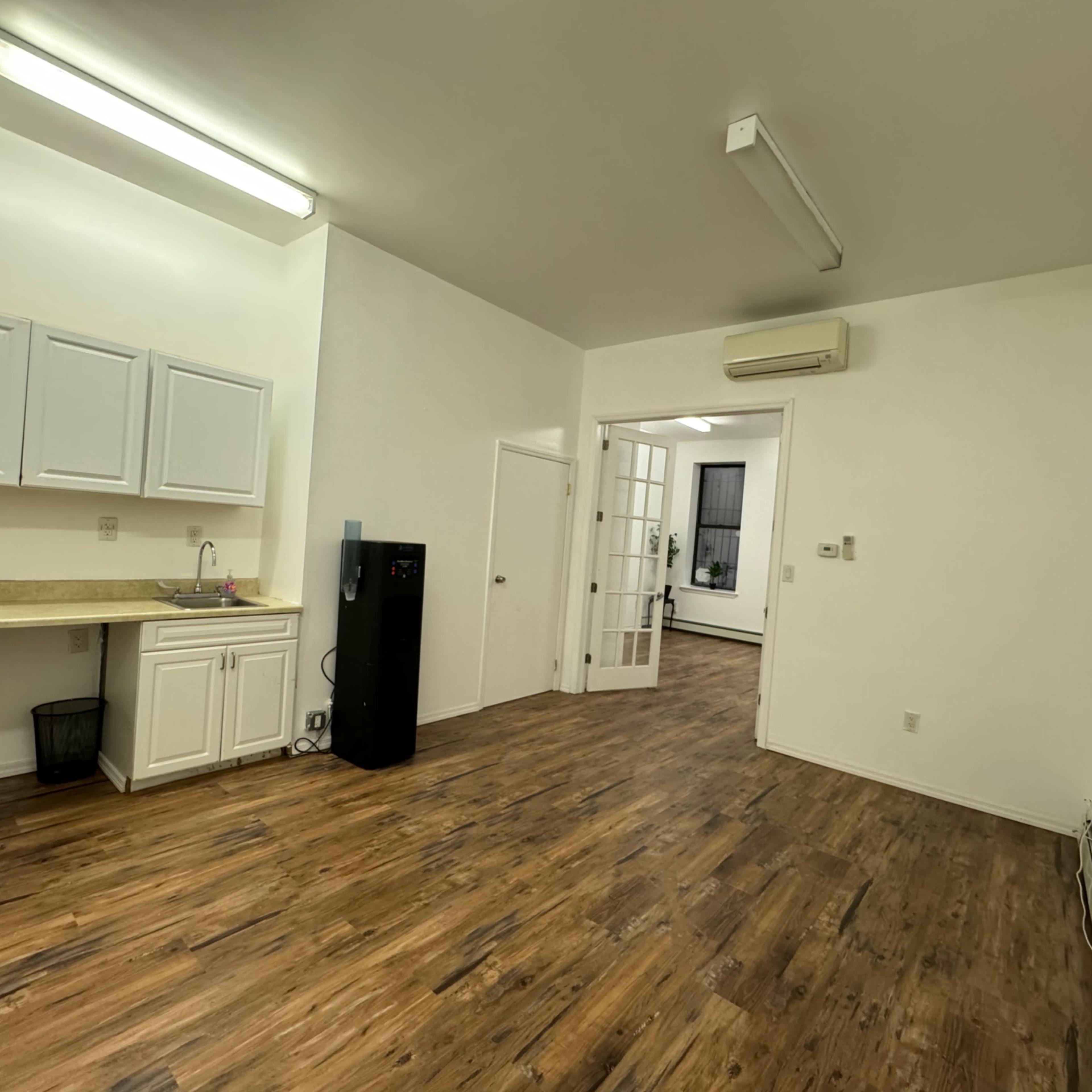 A spacious, empty room with a kitchenette on one side, featuring a refrigerator and a countertop, and an adjoining room visible through a set of glass doors.