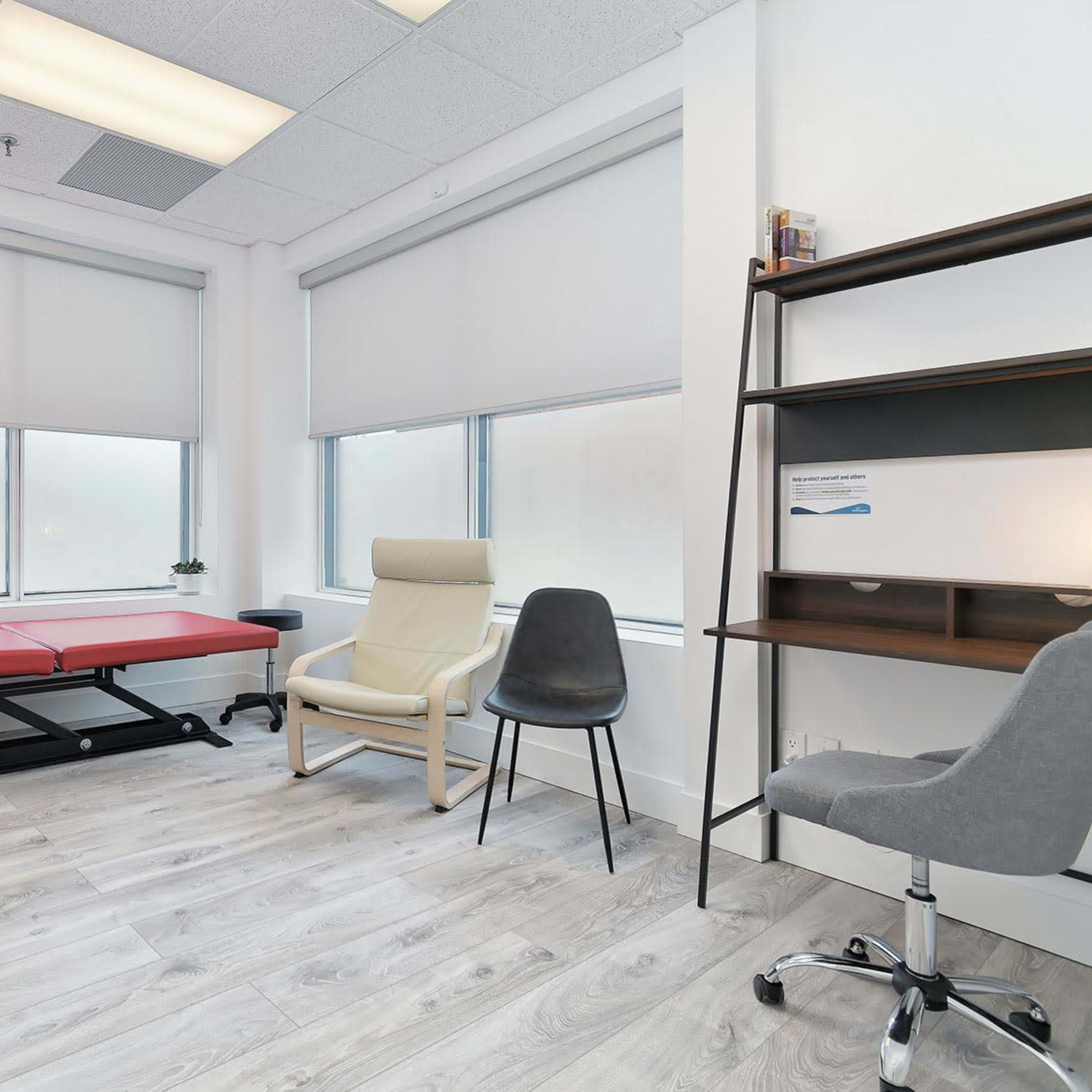 A minimally furnished therapy room with a massage table, a chair, and a small shelf, featuring large windows that allow natural light.