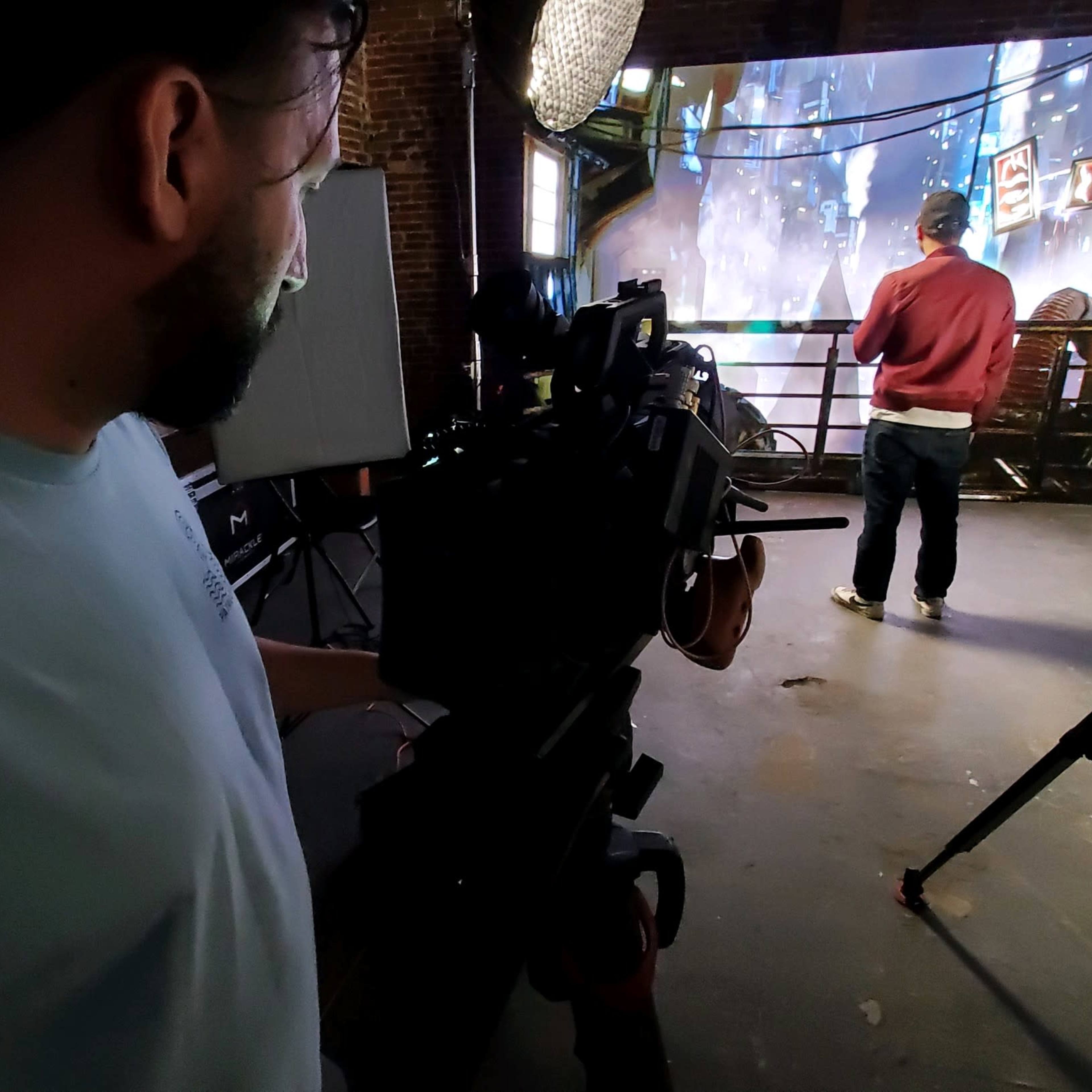 A person operates a camera while another individual stands in front of a projected urban scene on a wall of a studio.