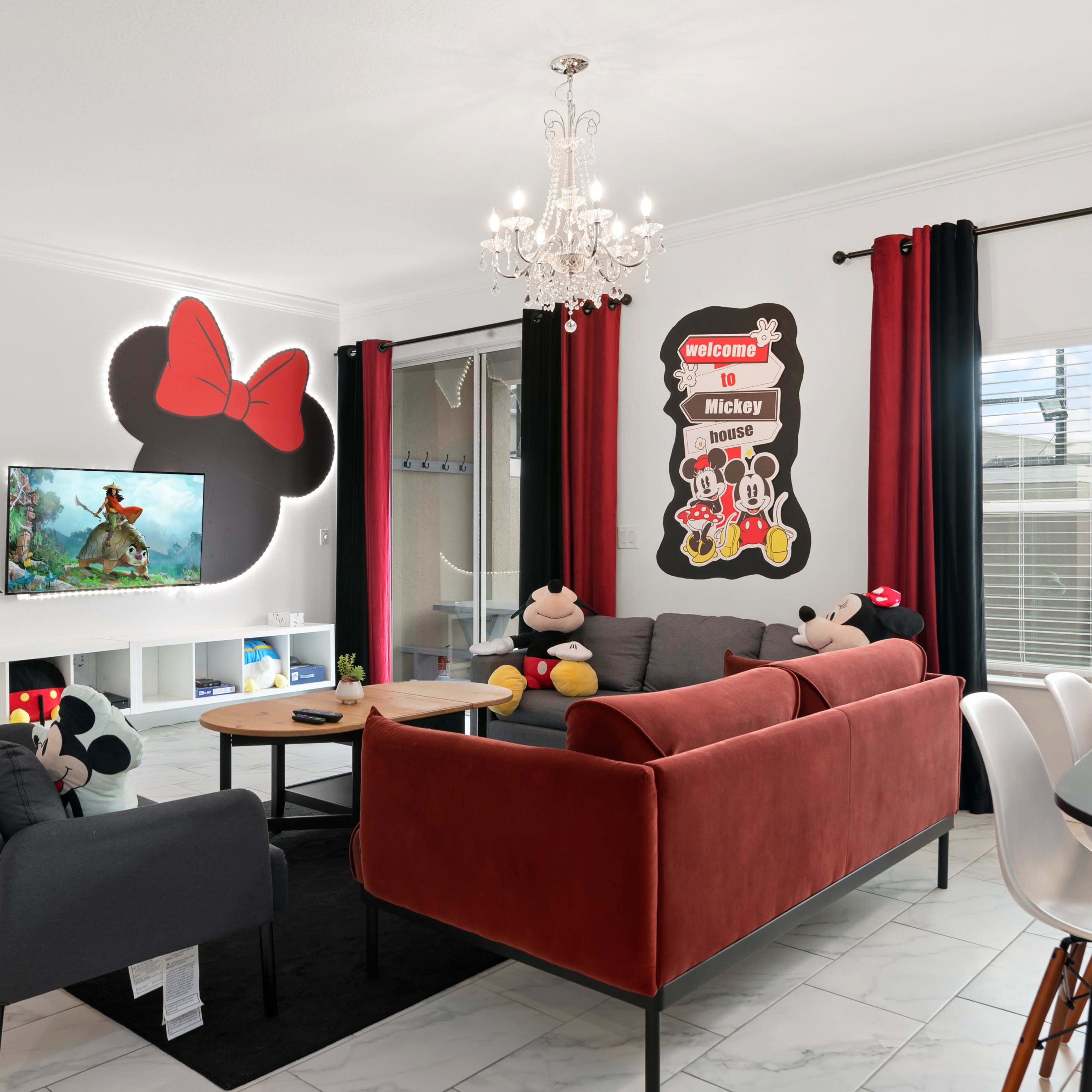 The living room features a mix of red and gray furniture, a wall decorated with Mickey Mouse art, and a chandelier above a seating area.