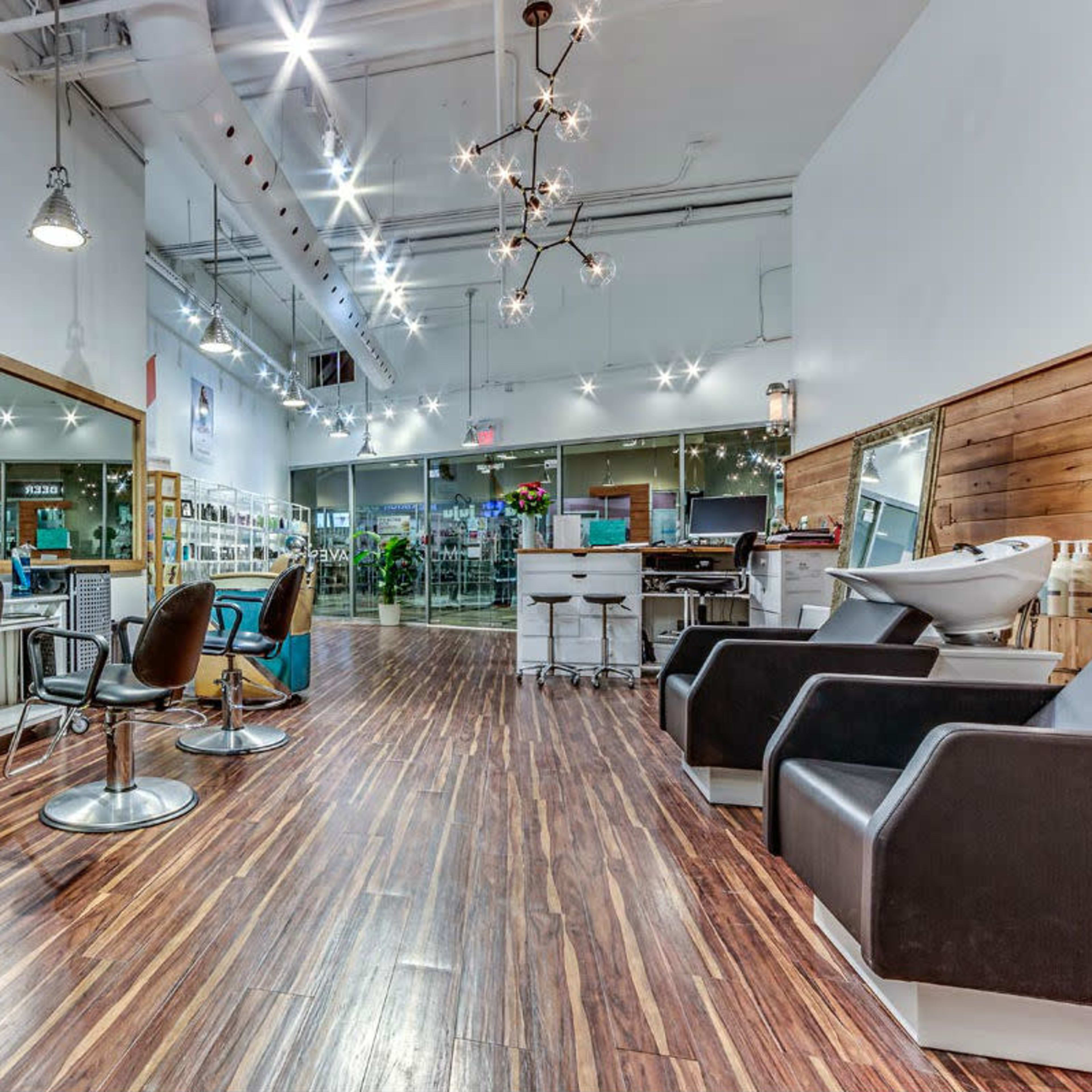 A modern hair salon with sleek wooden flooring, styling chairs, and a large mirror, illuminated by bright overhead lights.