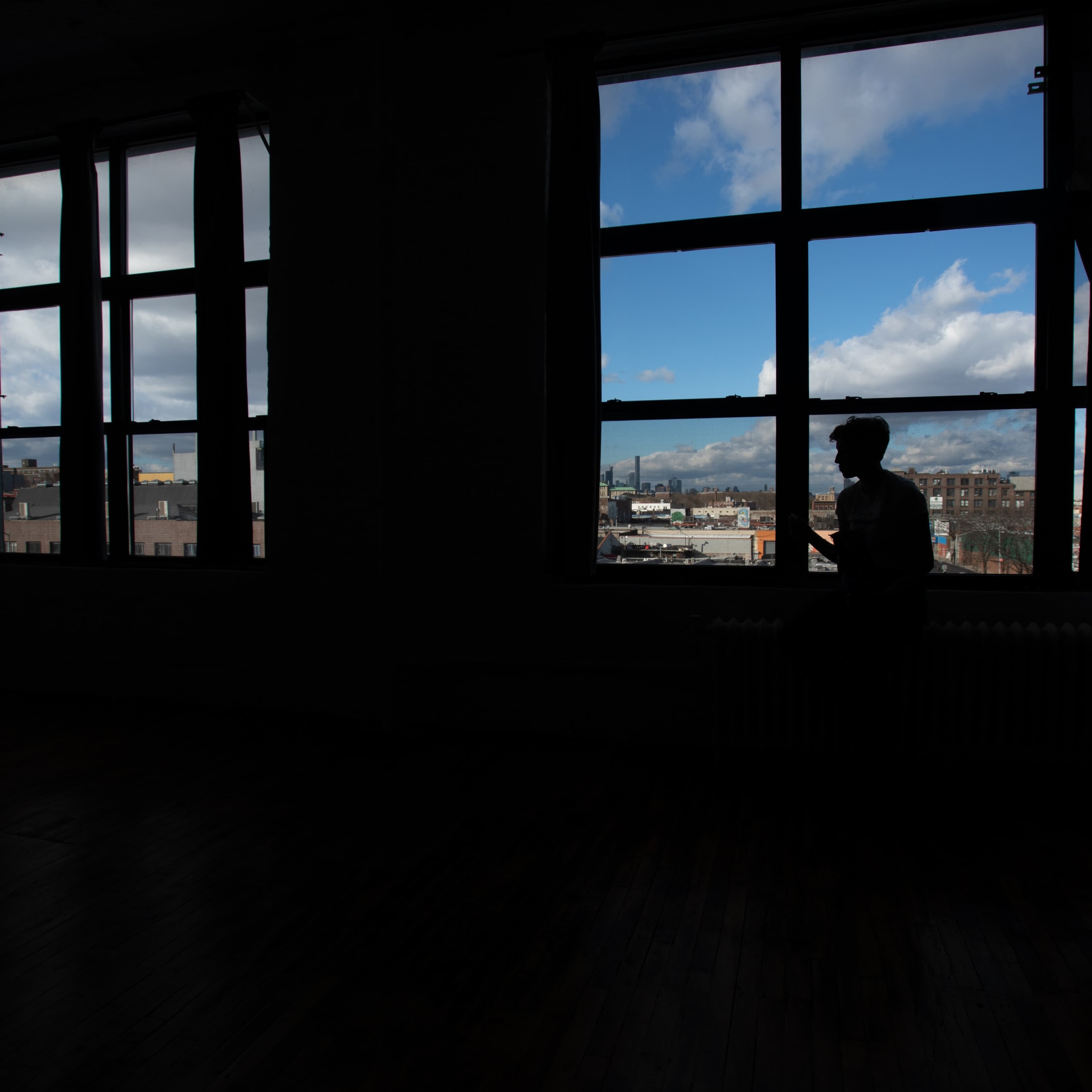 A person sits silhouetted in front of a large window, looking out at a cityscape under a partly cloudy sky.
