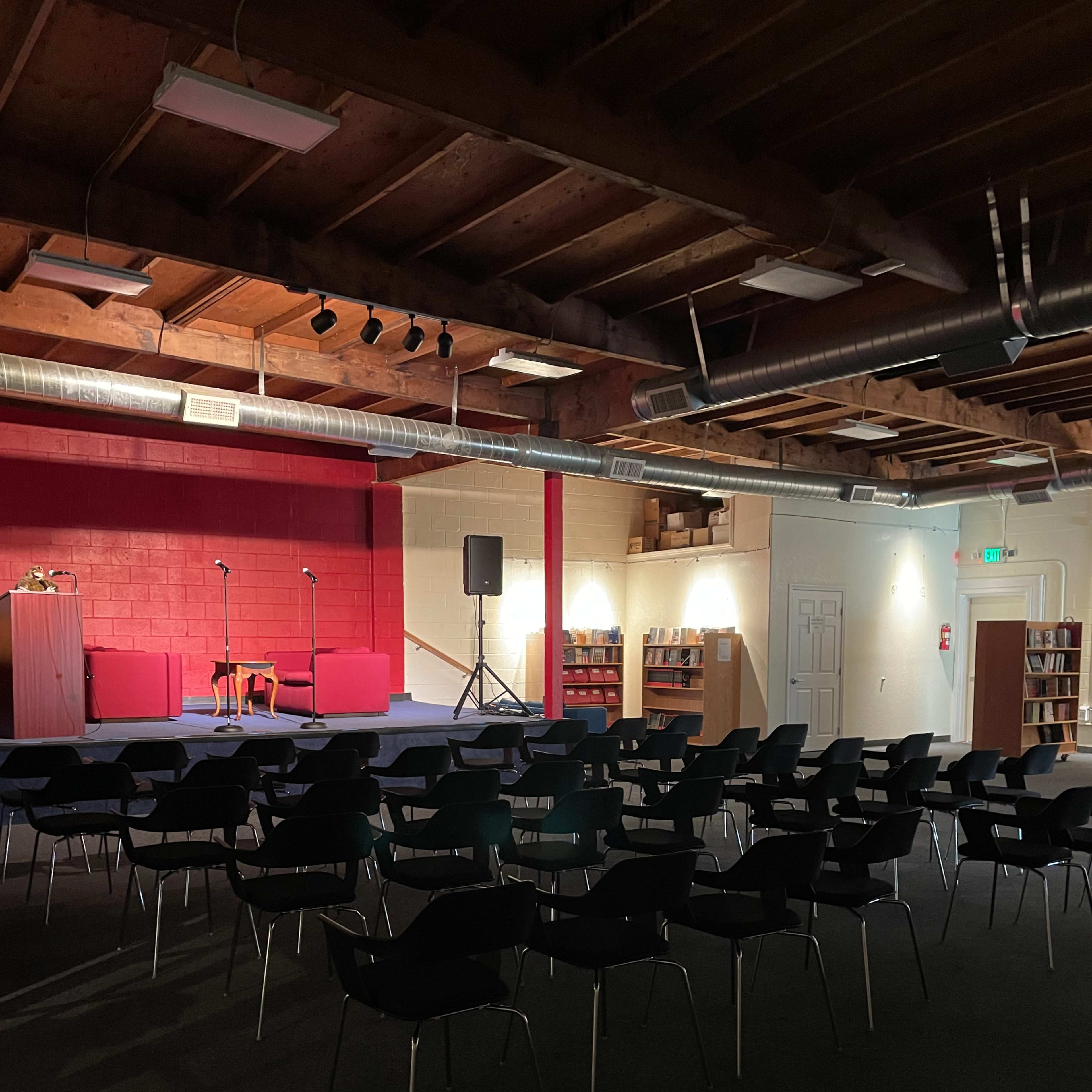 A dimly lit room with a small stage, two microphones on stands, and rows of black chairs facing the stage.