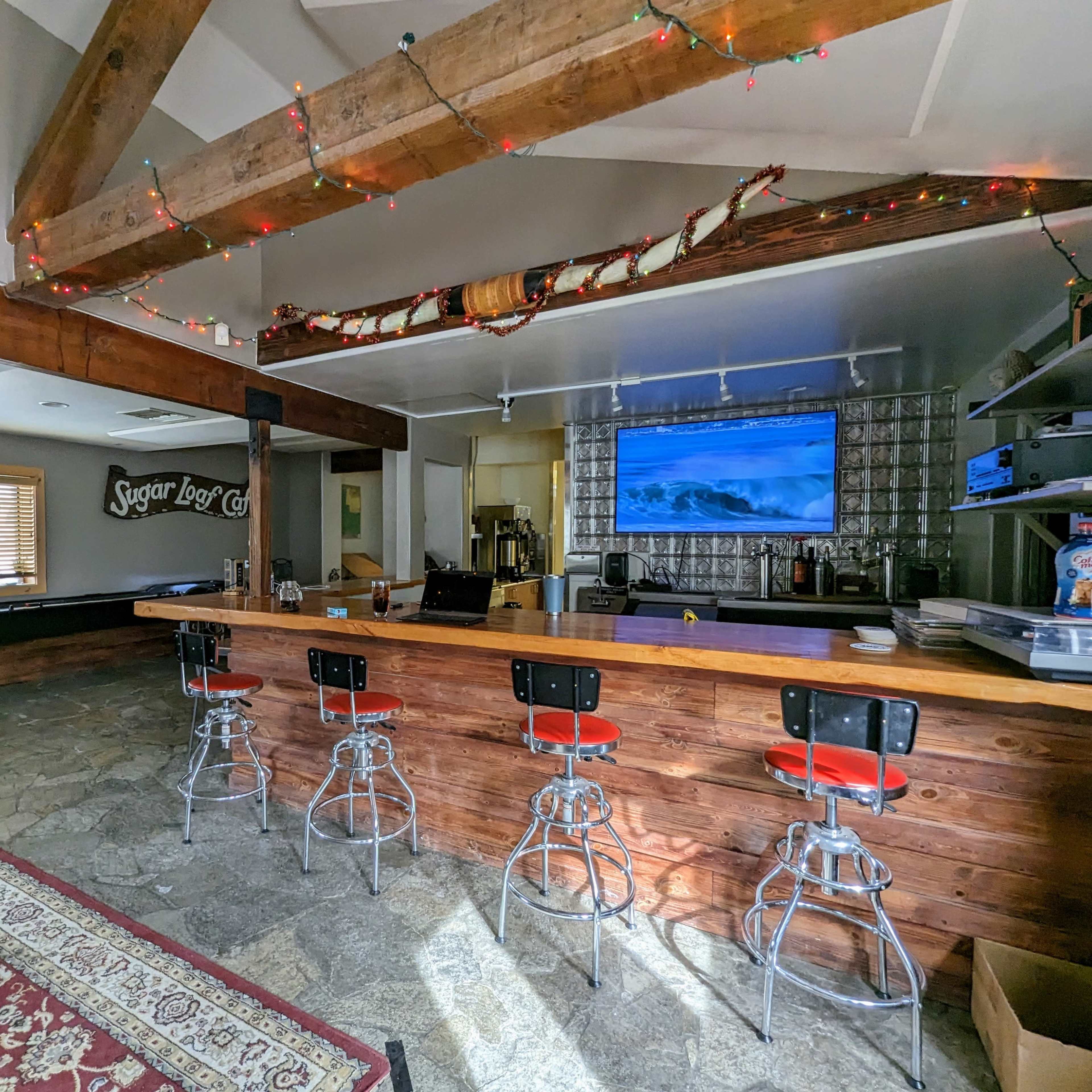A rustic bar area with wooden beams, a large television displaying ocean waves, and four black and red bar stools lined up along the counter.