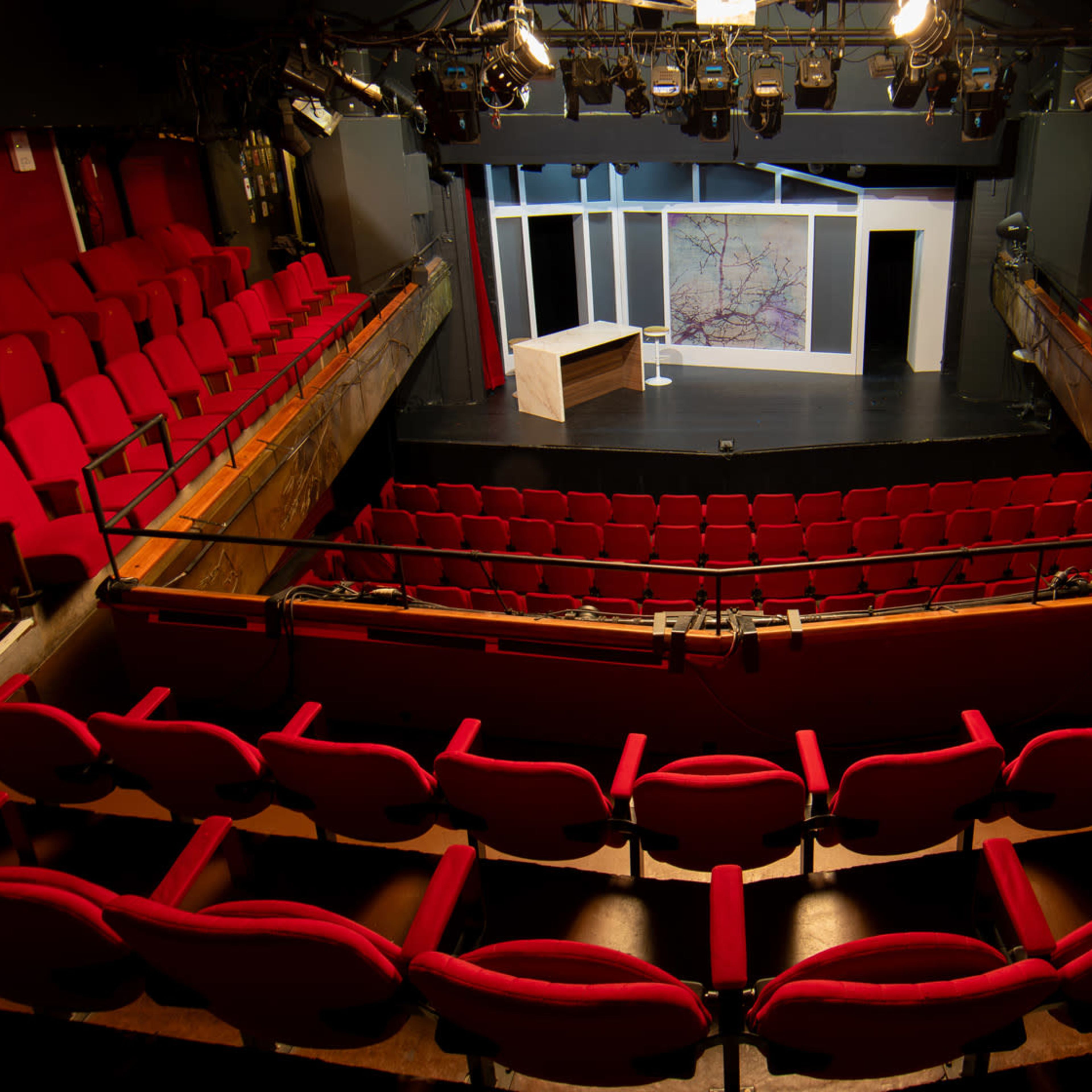 An empty theater interior features red plush seats arranged in a semicircle around a small stage with a simple set design.