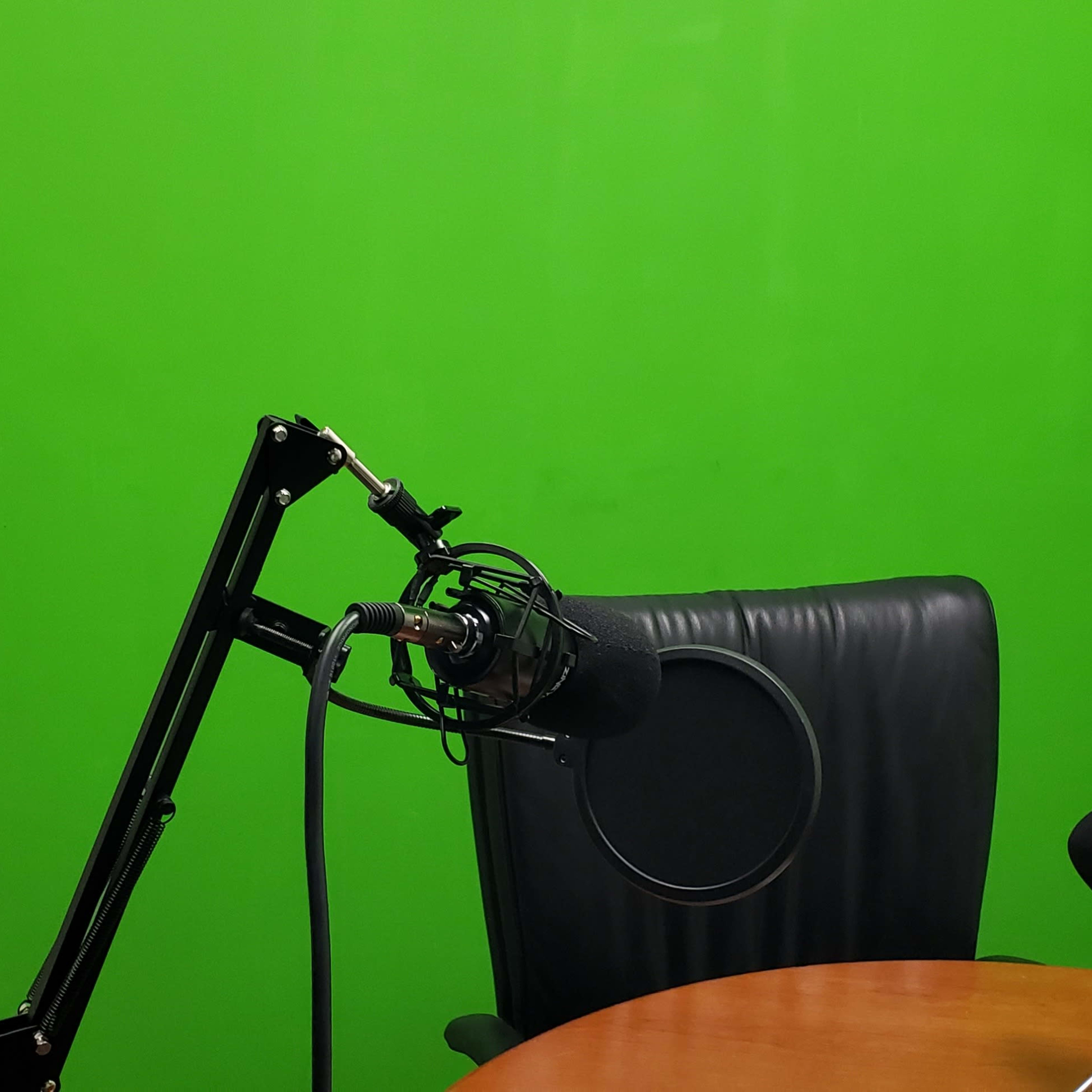 A black microphone mounted on an adjustable arm next to a chair and a wooden table in front of a green screen.