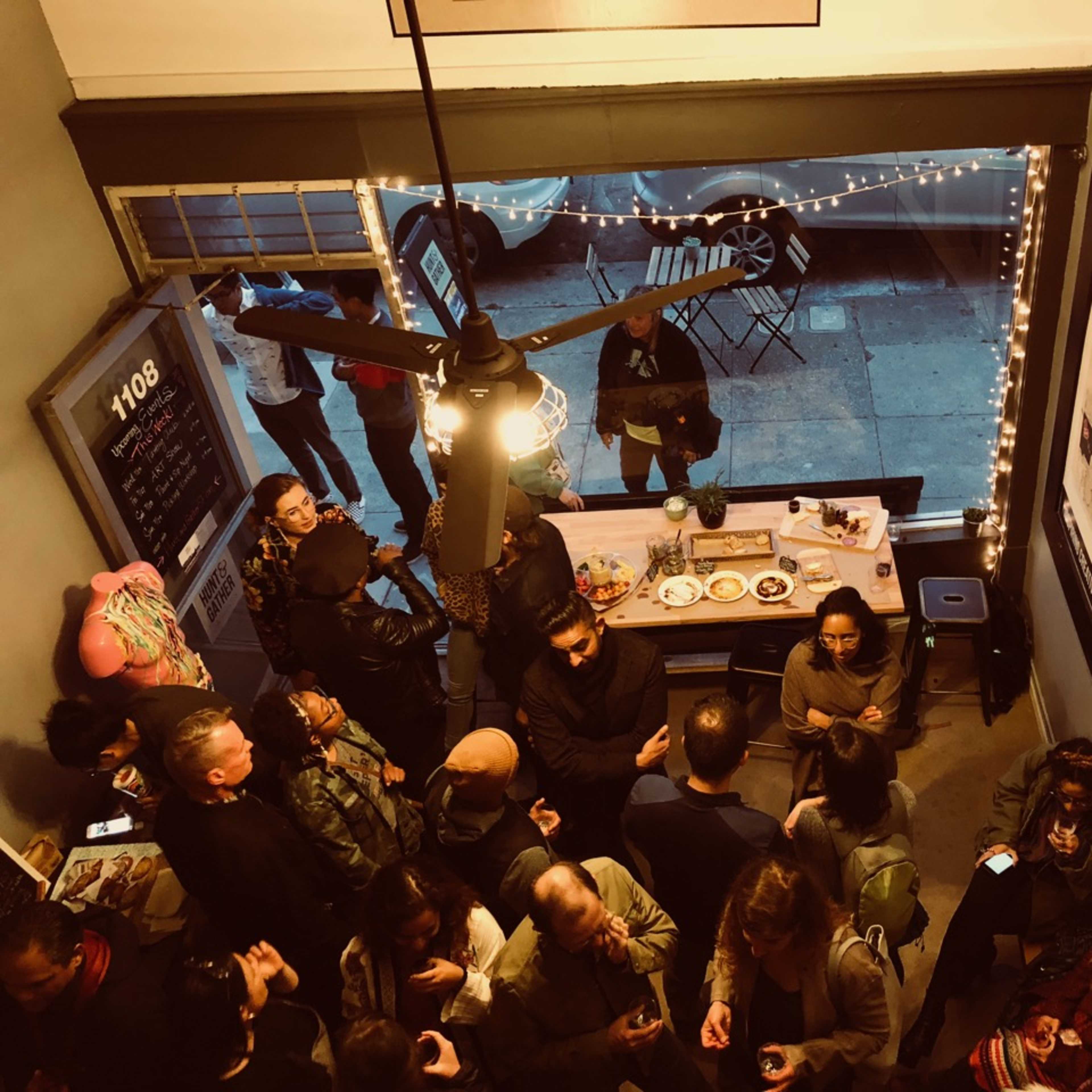 A crowded room with people socializing around a table filled with food and drinks, while art is displayed on the walls and lights hang above.