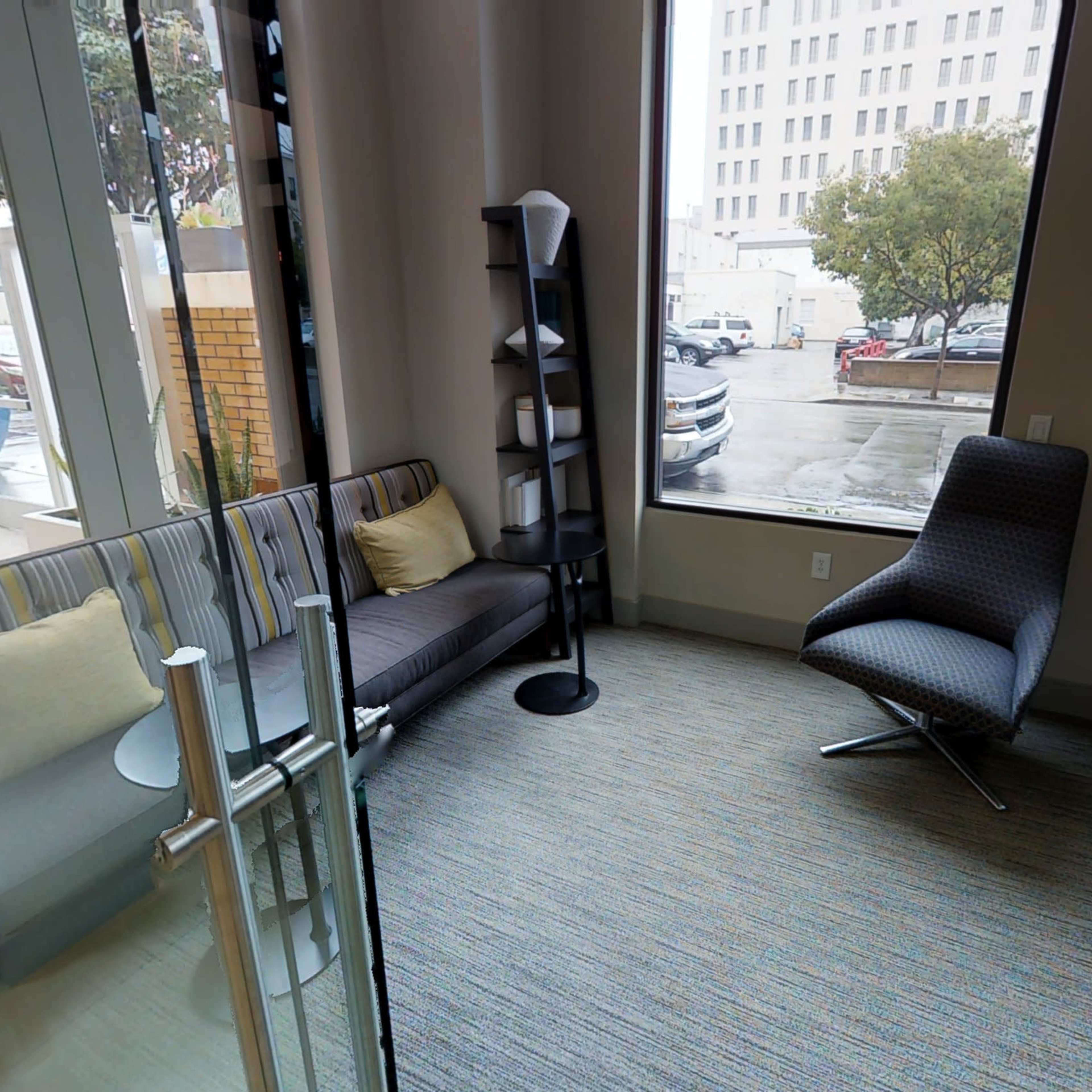A small lounge area with a couch, a single chair, and a bookshelf, all situated next to large windows.