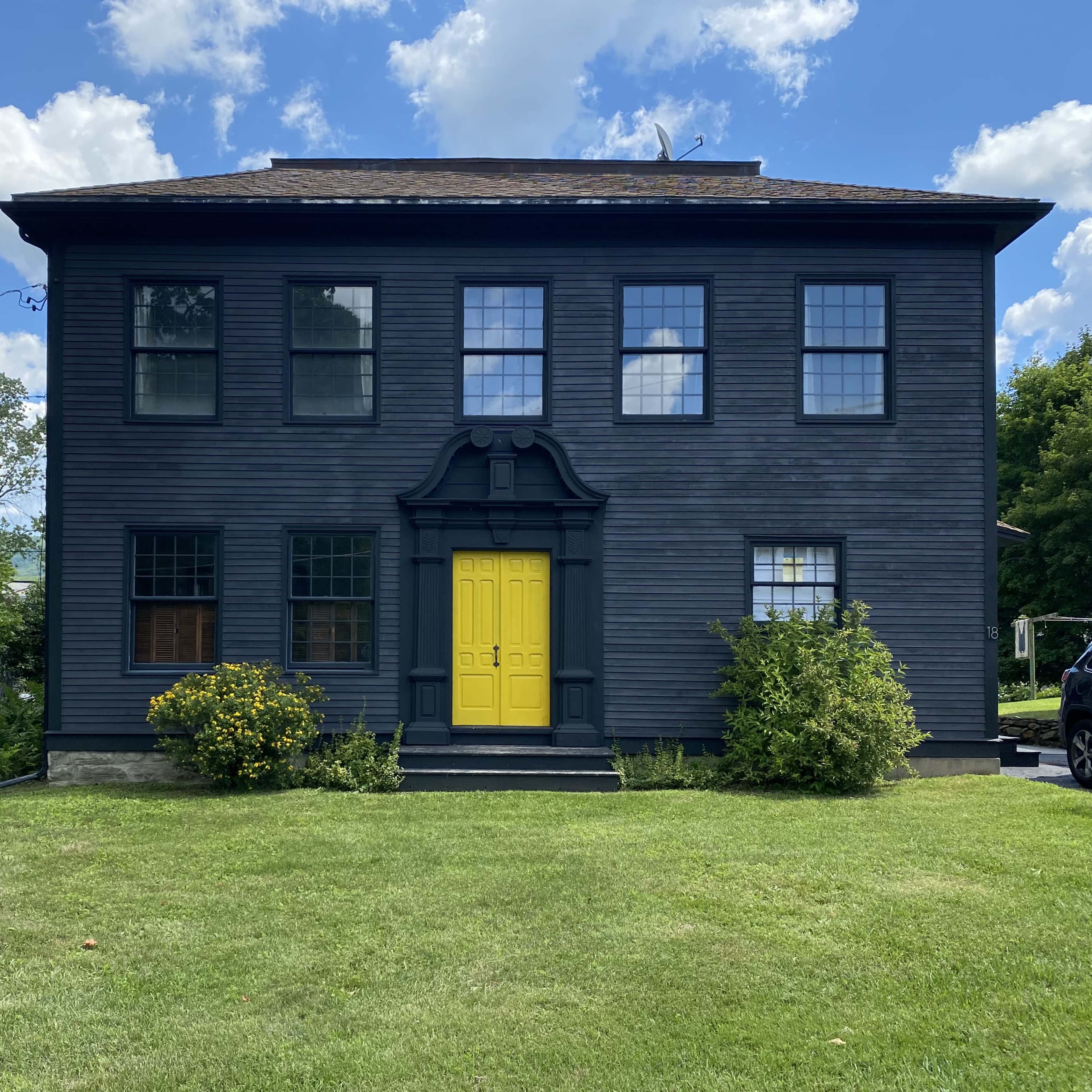 A two-story black house with a bright yellow front door and multiple windows, situated on a grassy lot with shrubs and trees nearby.