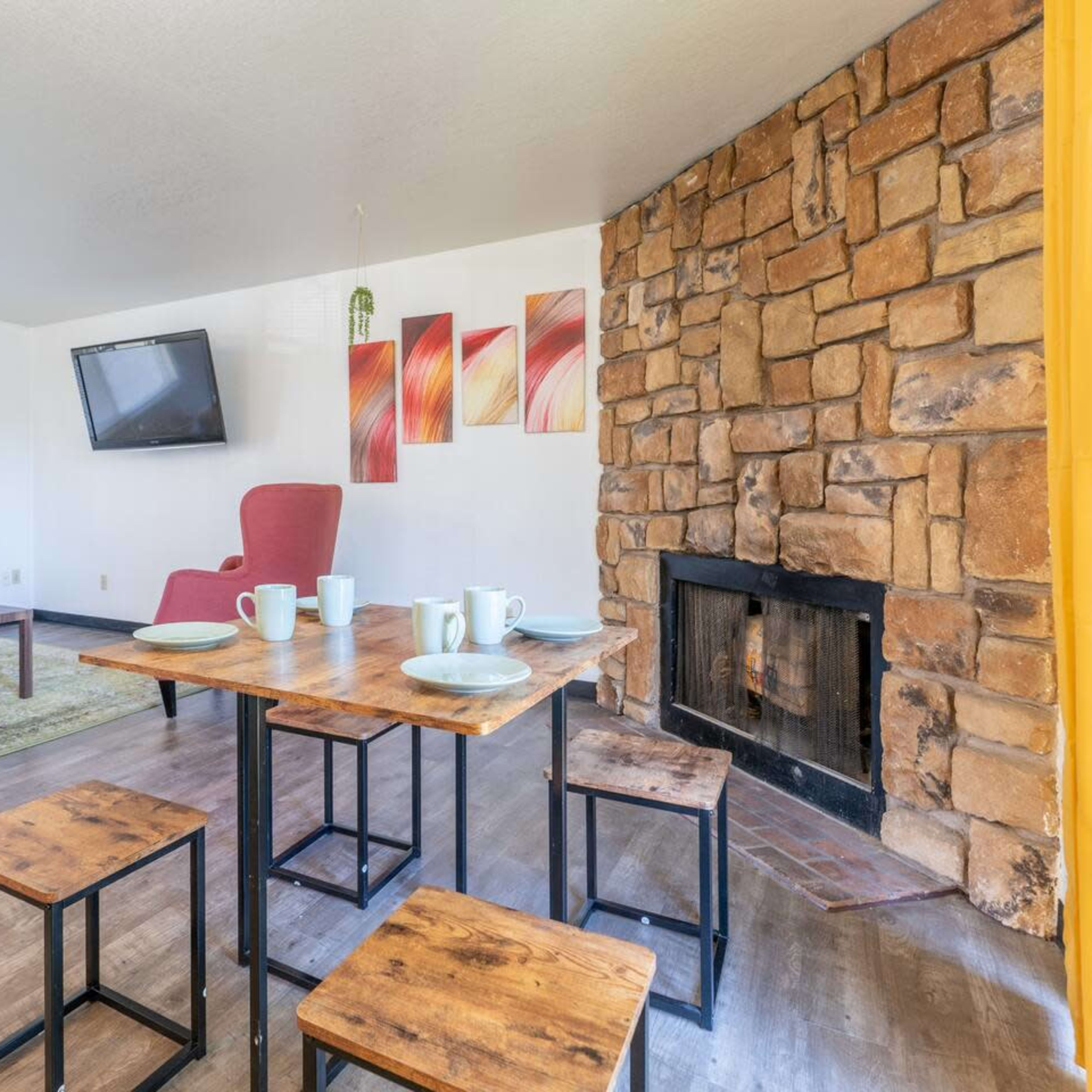 A cozy living space with a stone fireplace, a wooden dining table set for four, a television mounted on the wall, and a red armchair.