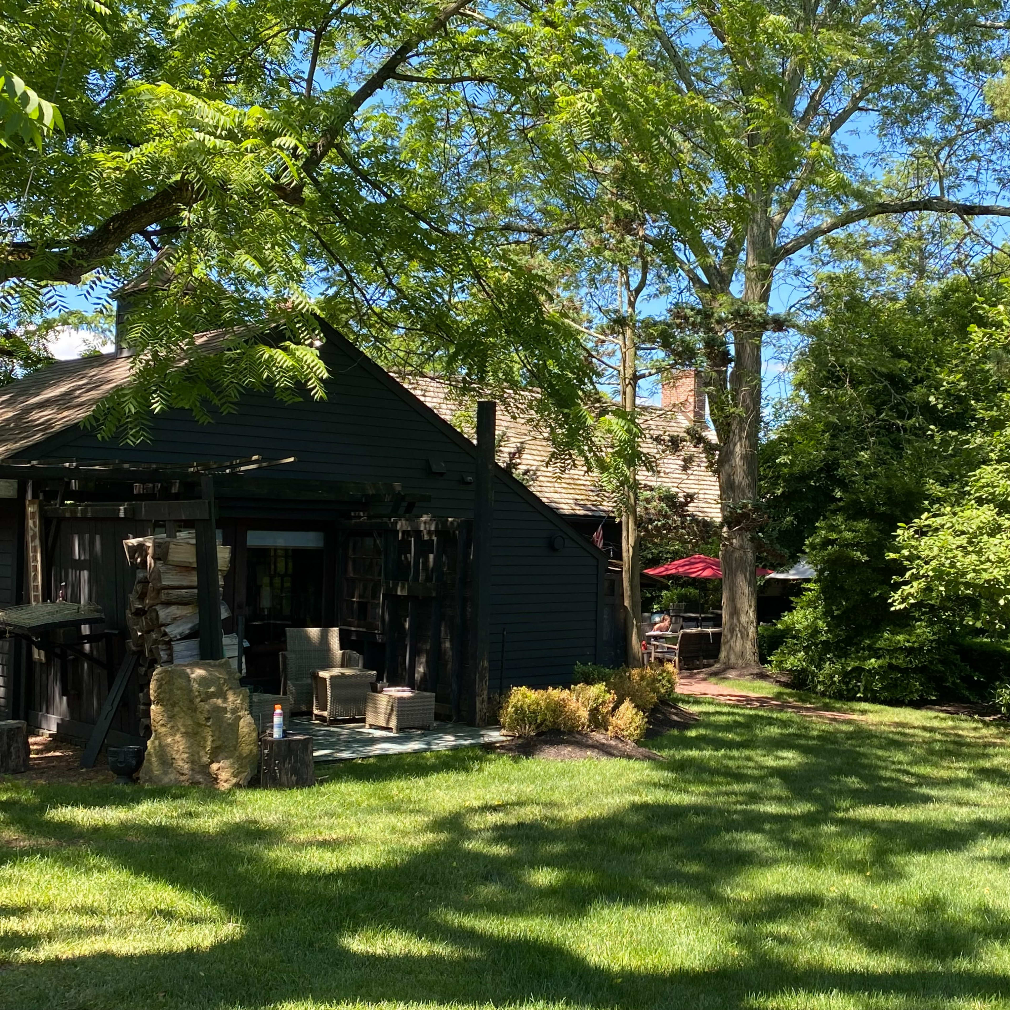 A rustic black wooden building surrounded by lush green grass and trees, with a hint of red from an outdoor umbrella visible in the background.