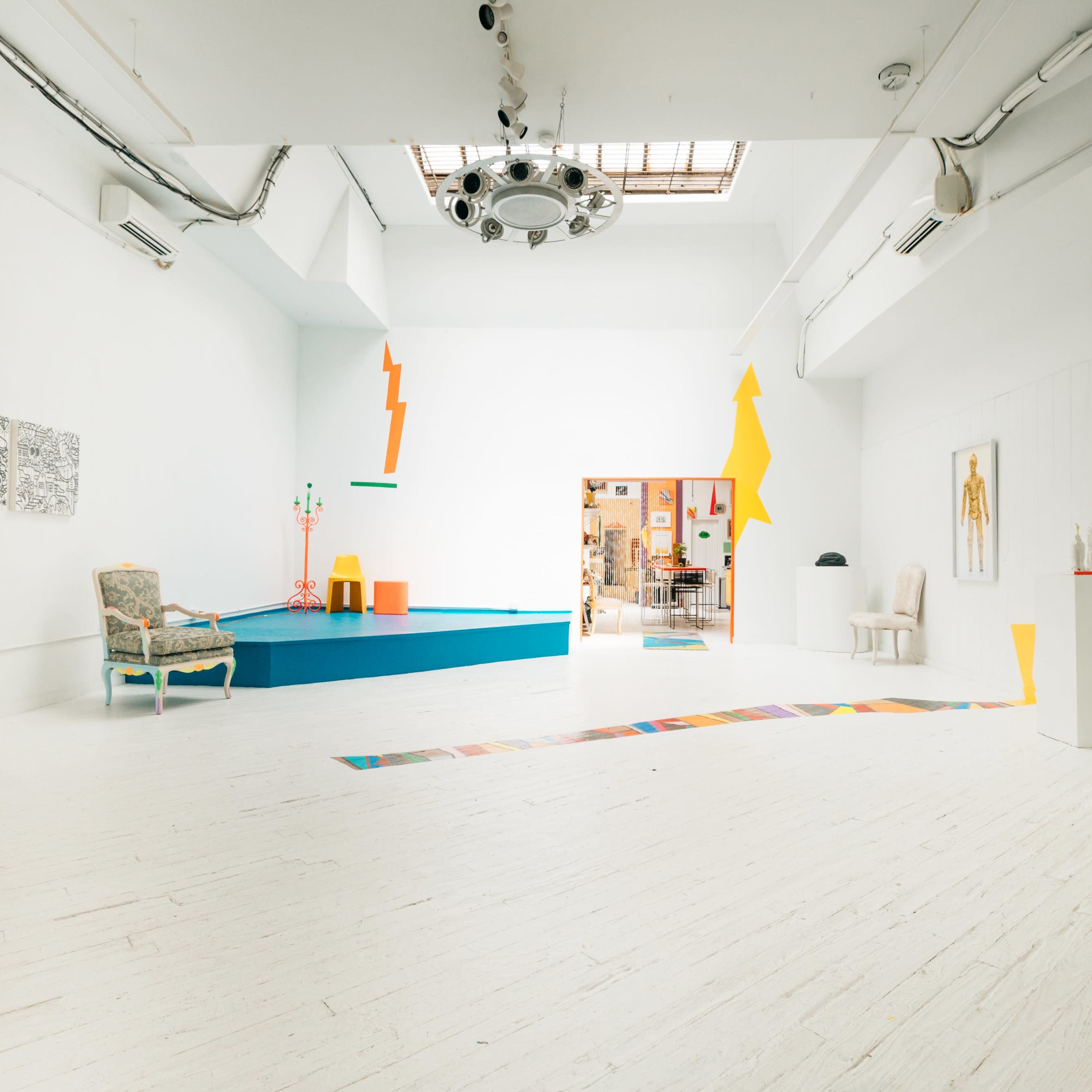 A bright, spacious gallery with white walls, colorful geometric decorations, and a few pieces of furniture arranged around a vibrant blue platform.