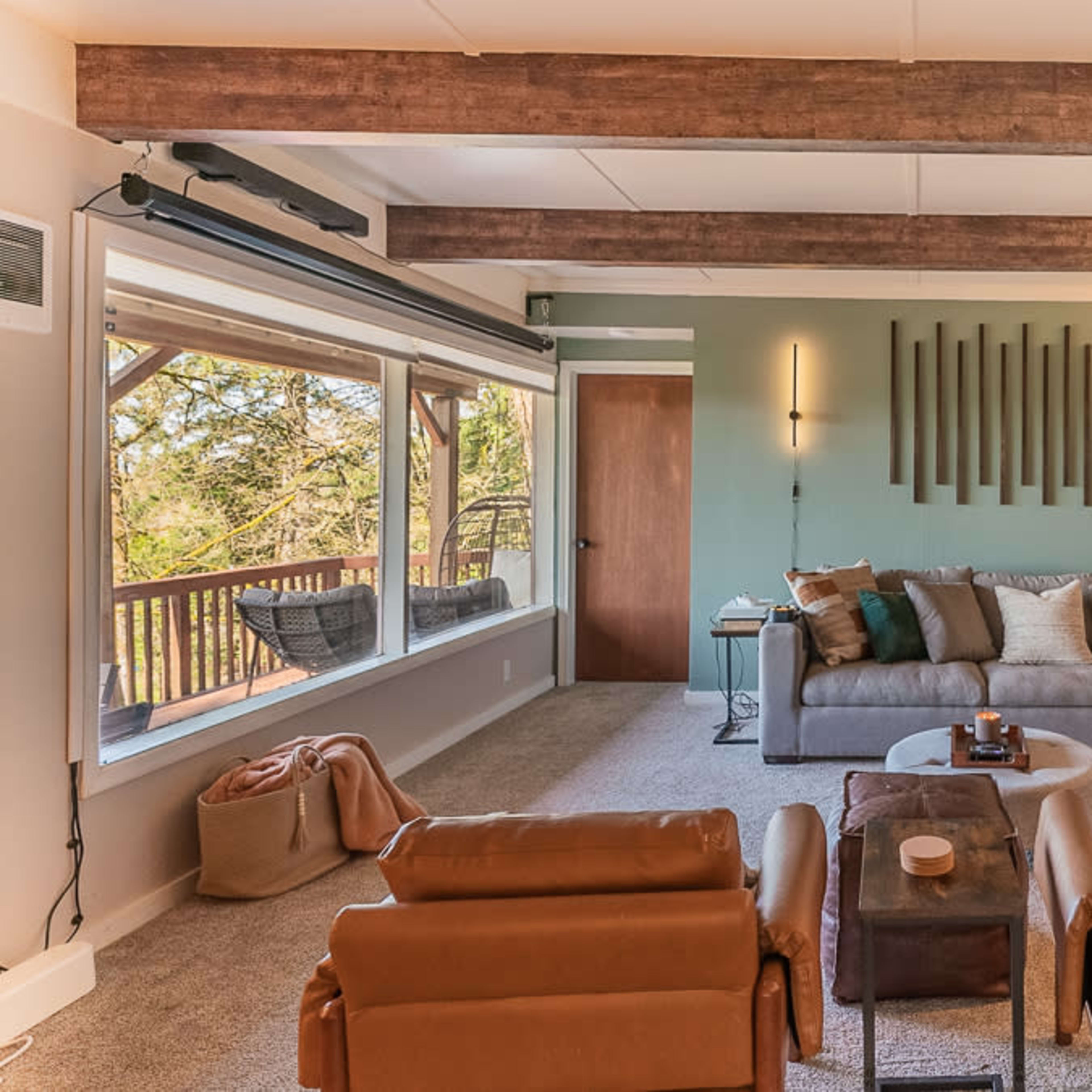 A modern living room features a large sectional sofa, two leather chairs, and a window that opens to a forest view.
