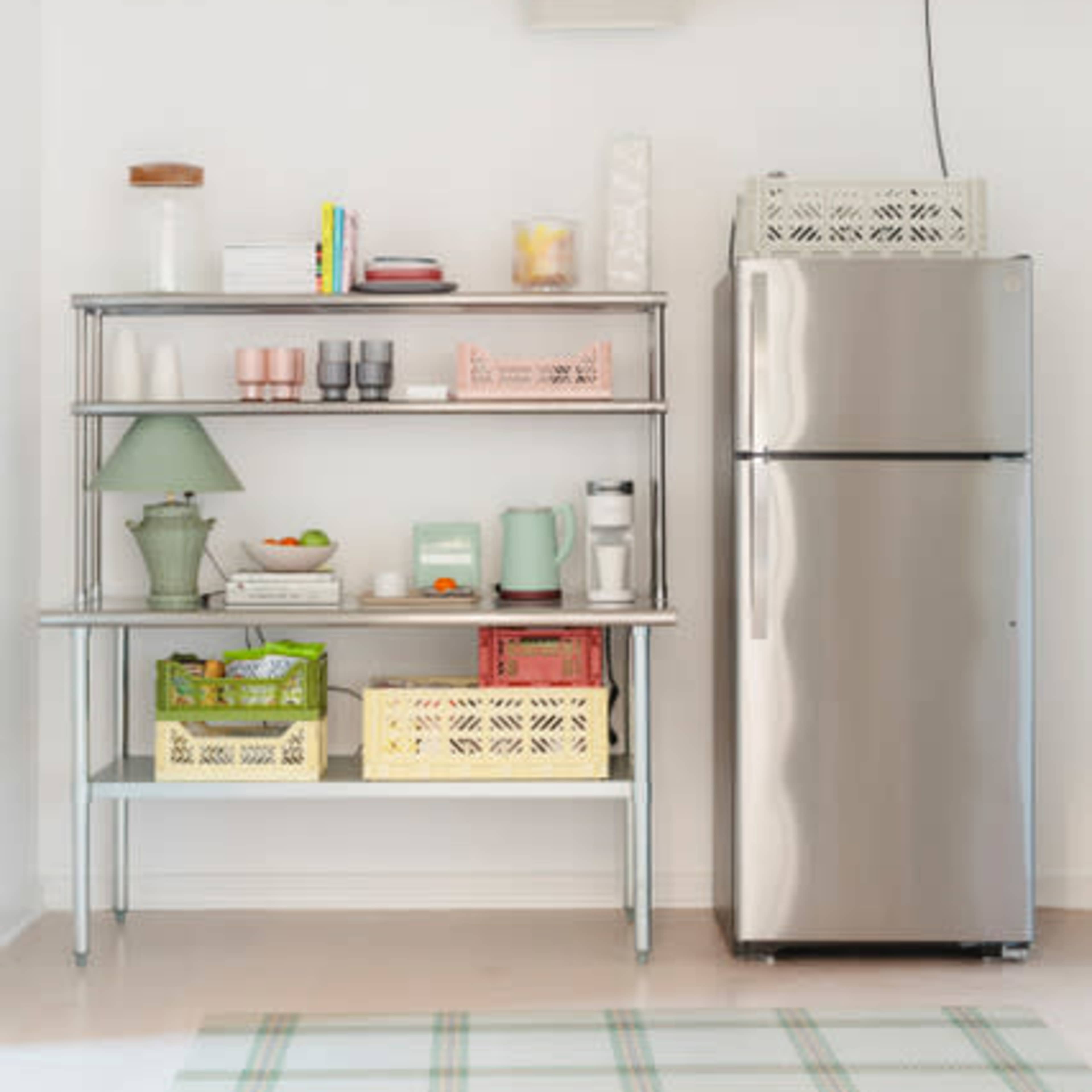 A metal shelving unit with various kitchen items sits next to a stainless steel refrigerator in a bright, open space.