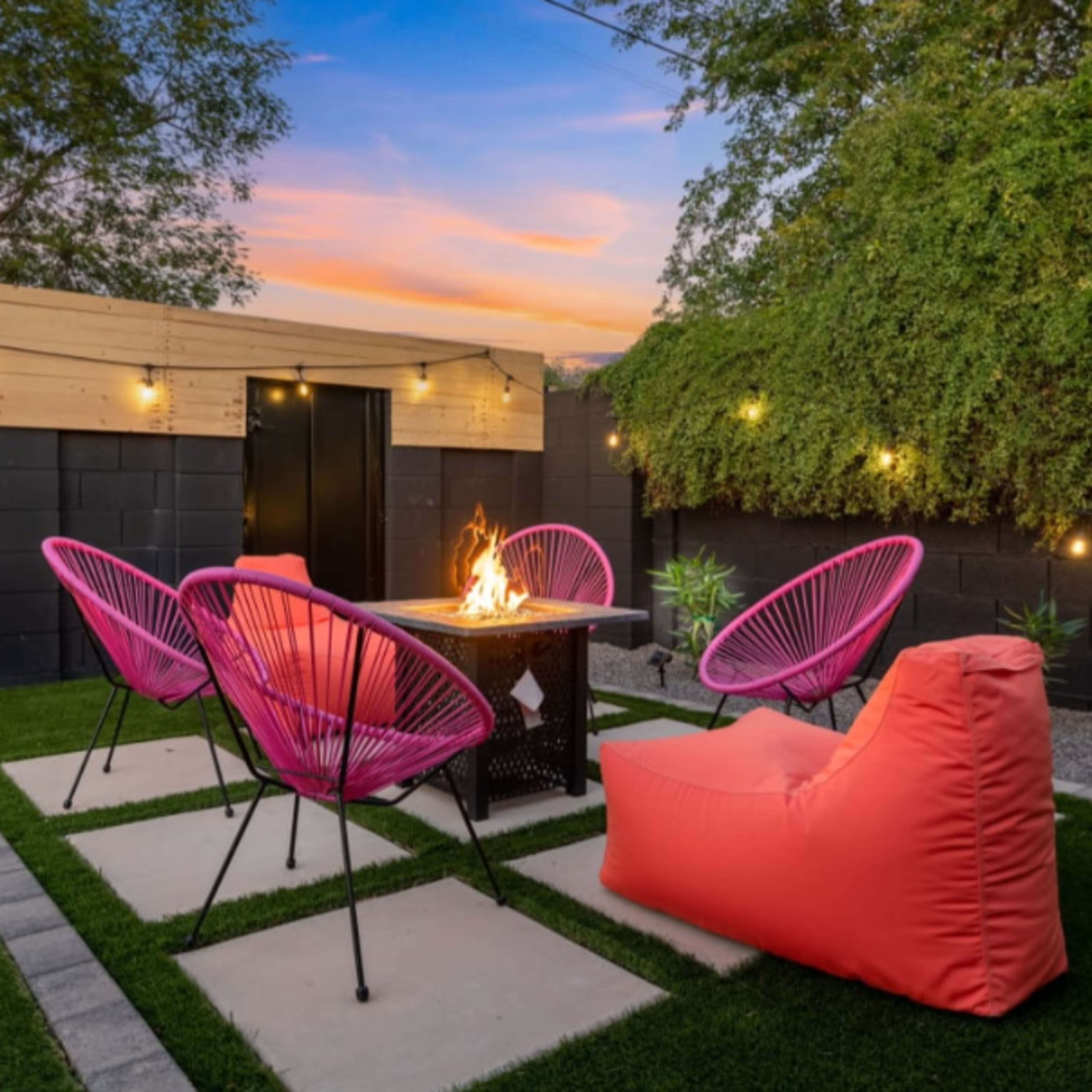 A cozy outdoor seating area with four pink chairs and a fire pit surrounded by a grassy lawn and adorned with string lights.