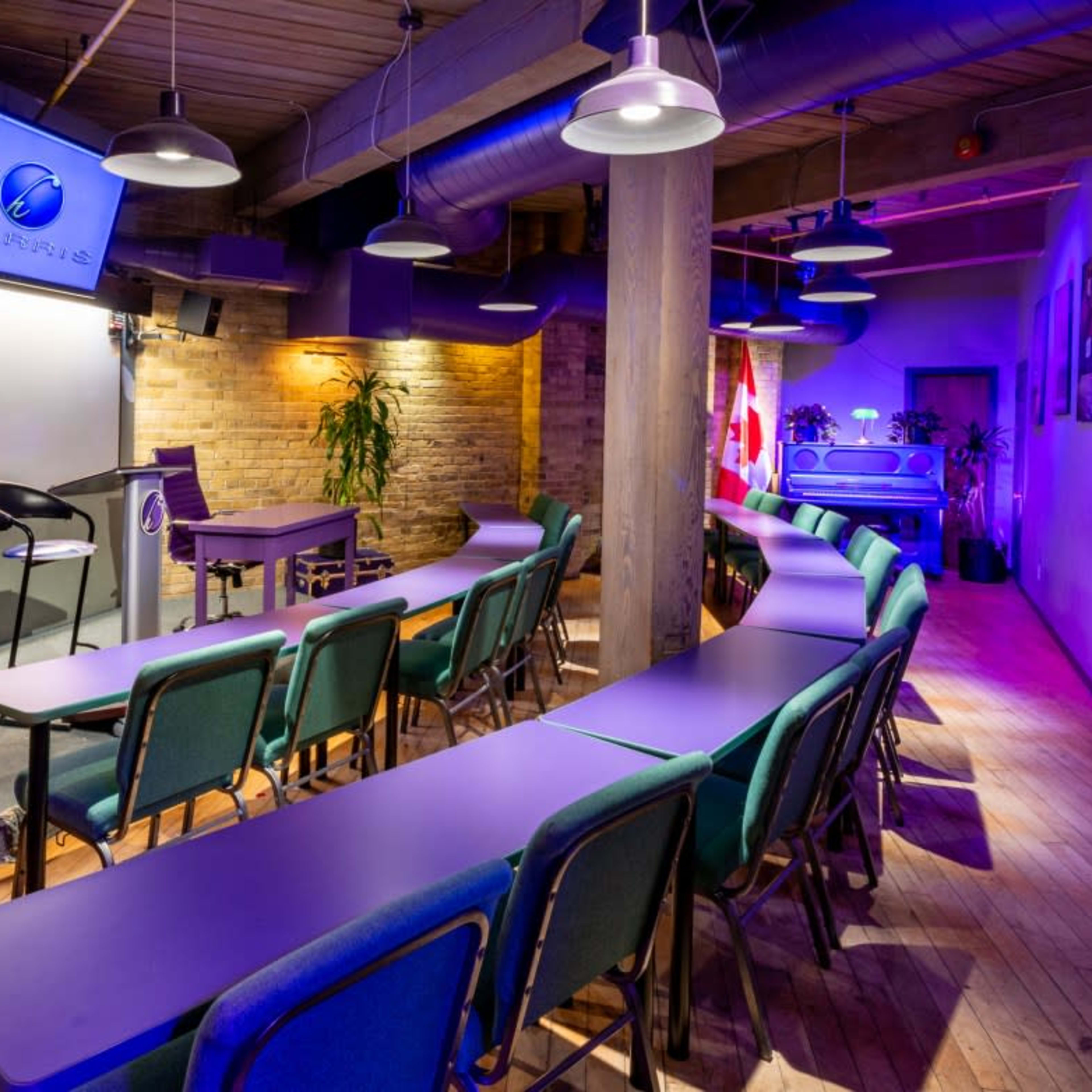 A seminar room with rows of purple tables and chairs, a projector screen, and plants, illuminated by purple and blue lighting.