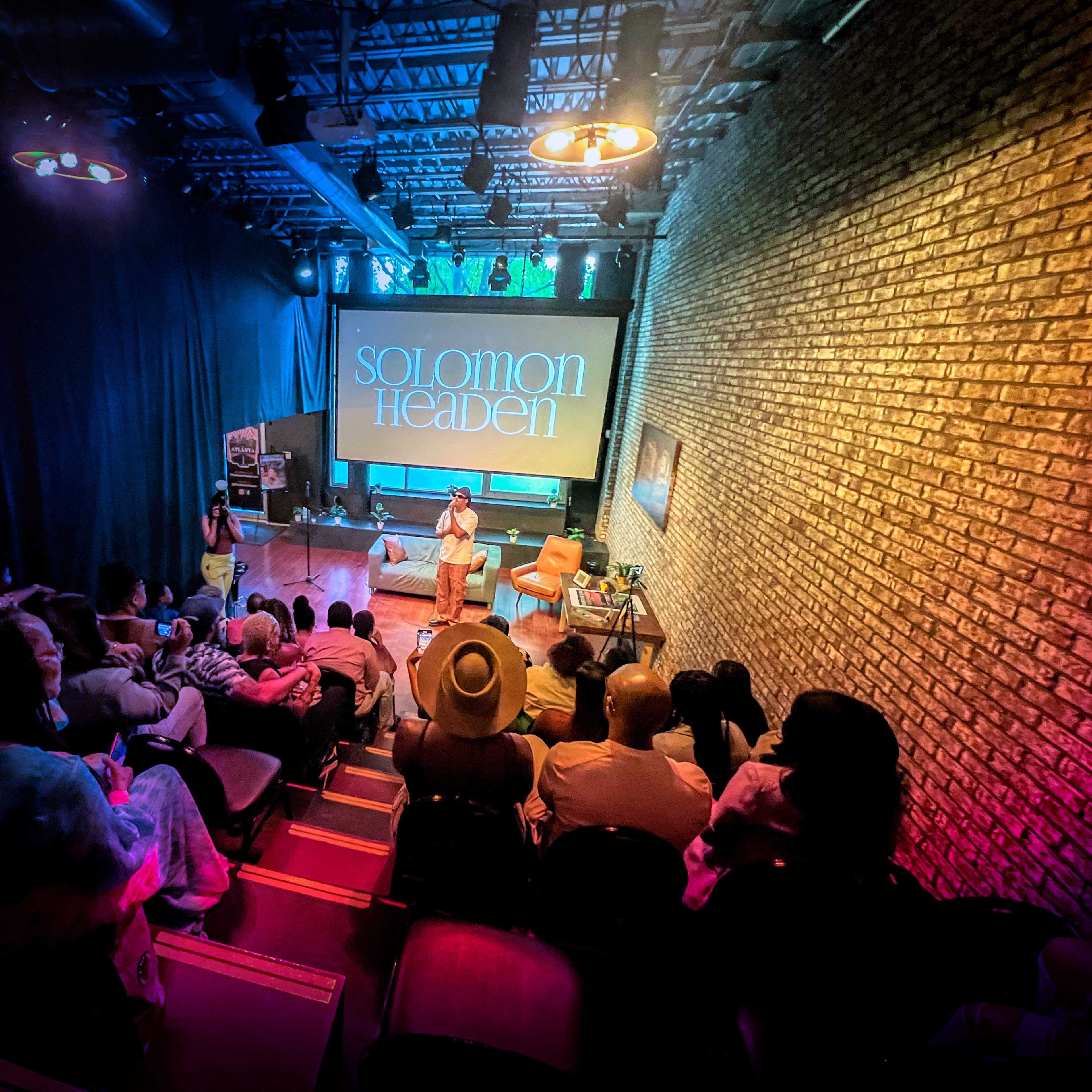 A speaker presents on stage at an event in an intimate venue, with an audience seated in rows before a screen displaying "SOLOMON HEADEN."