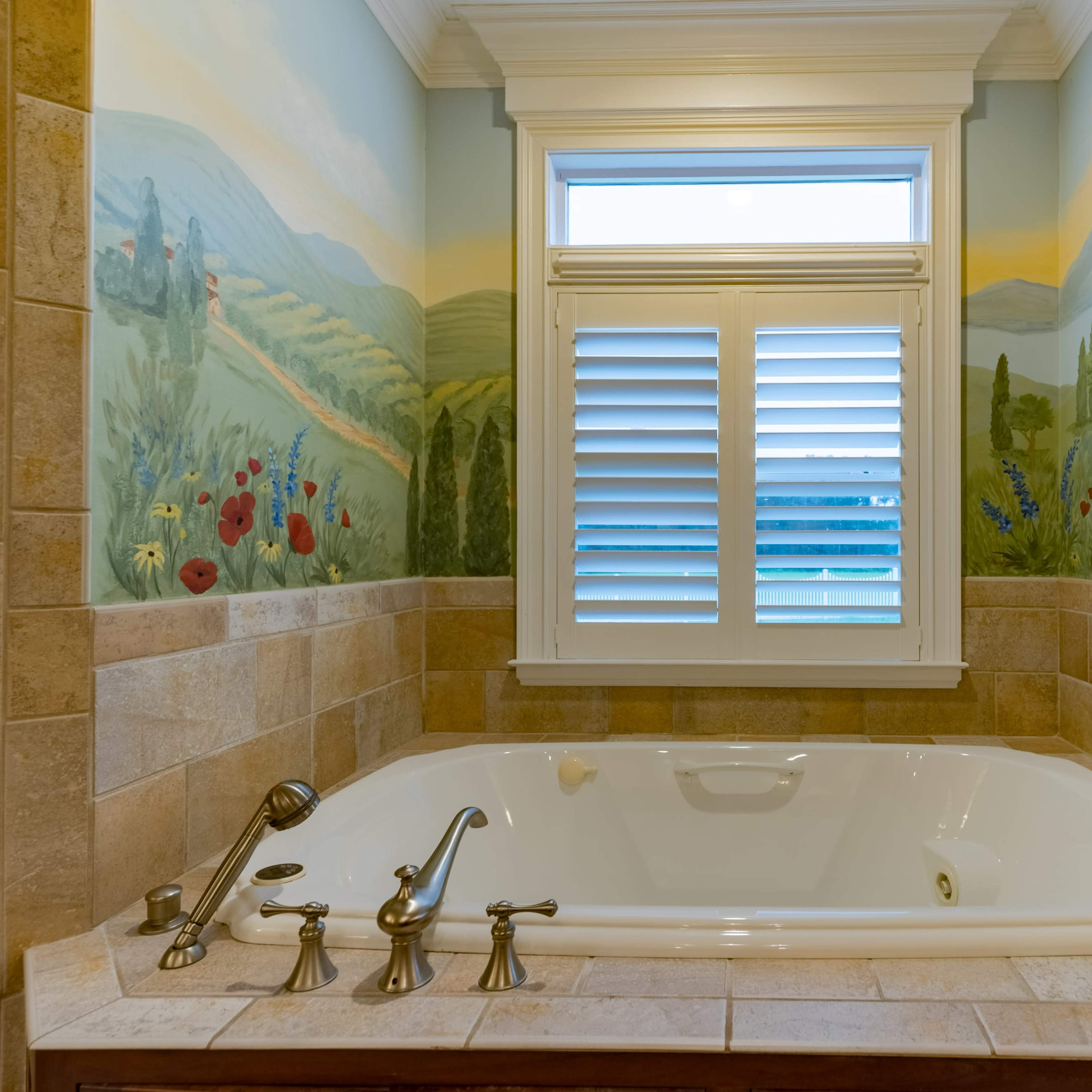 A jetted bathtub is positioned against a wall adorned with a mural of rolling hills and flowers, with a window above letting in natural light.